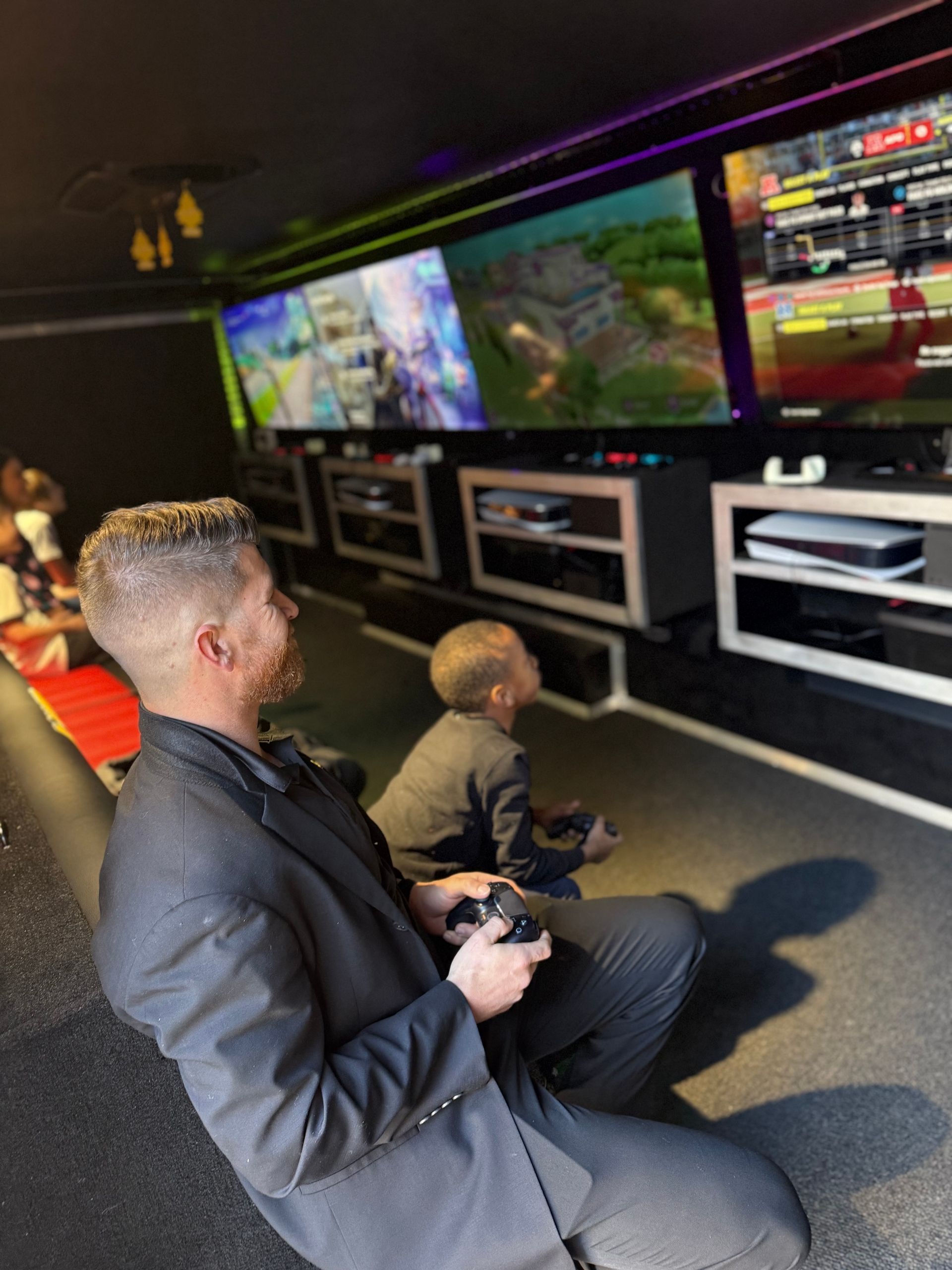 An adult and a child in formal wear playing video games on multiple large monitors in a darkened, stadium-style room.