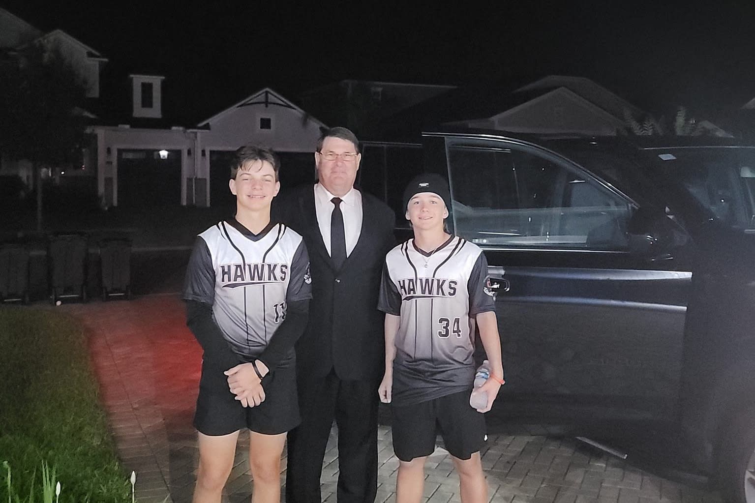Three people posing outdoors at night beside a black vehicle, one in a suit and two in sports uniforms.