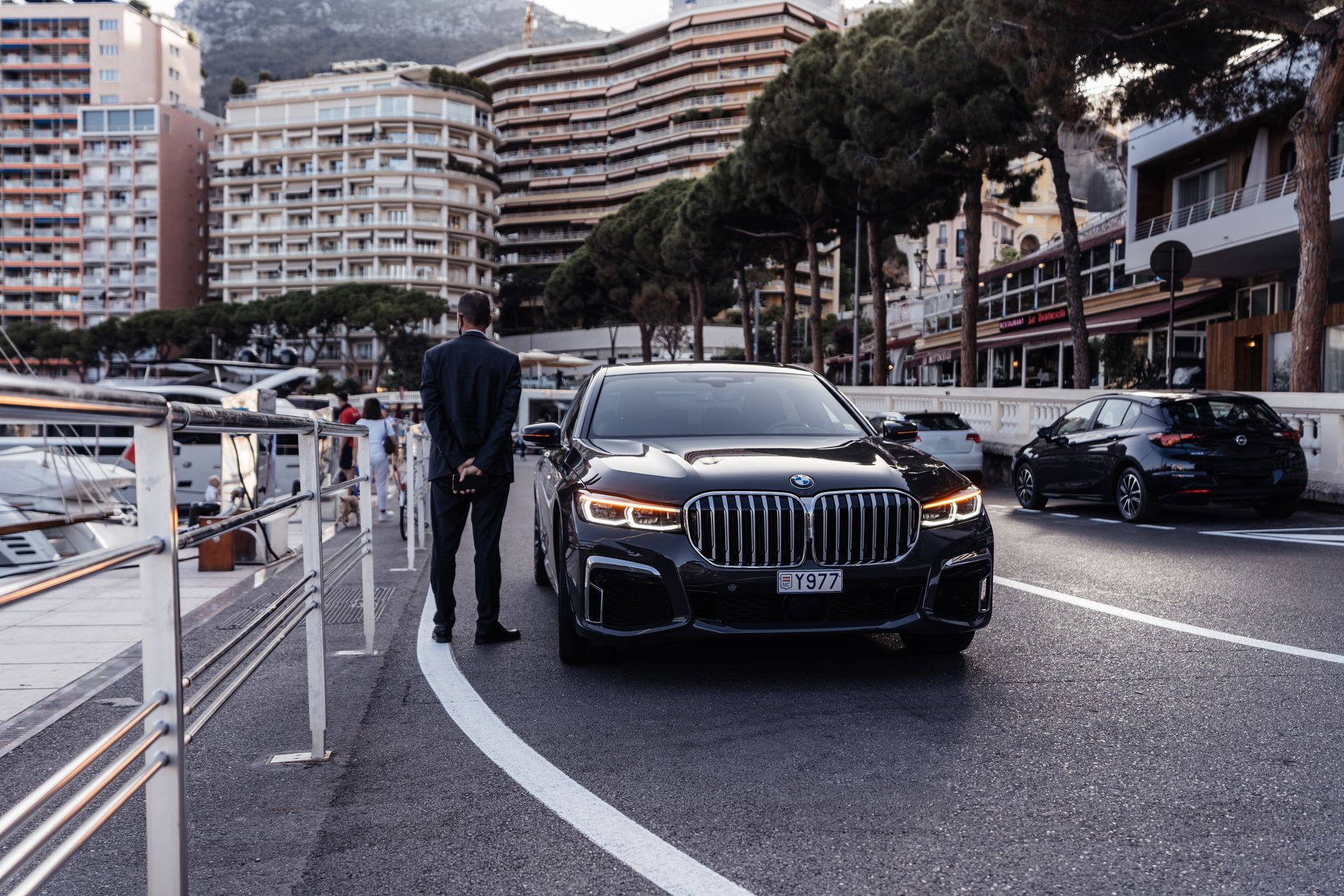Black luxury SUV parked on a waterfront road beside a person, with city buildings in the background