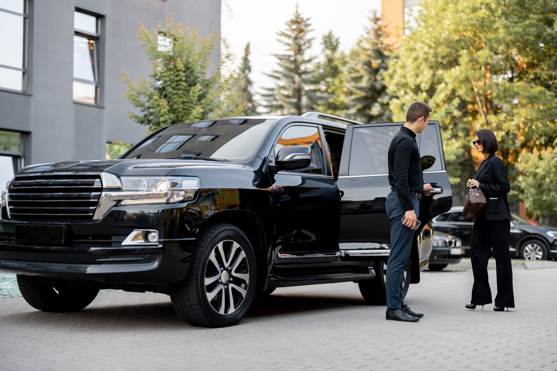 Black SUV parked outside a building with two people standing nearby, one by the open rear door