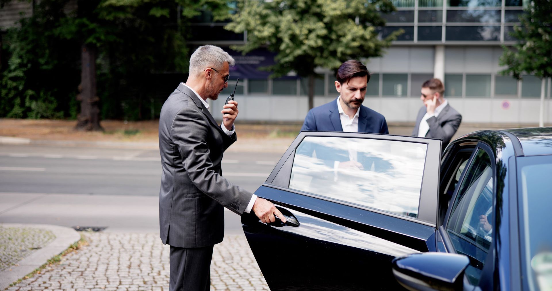 Man in a suit opening a car door while two people stand nearby on a city street