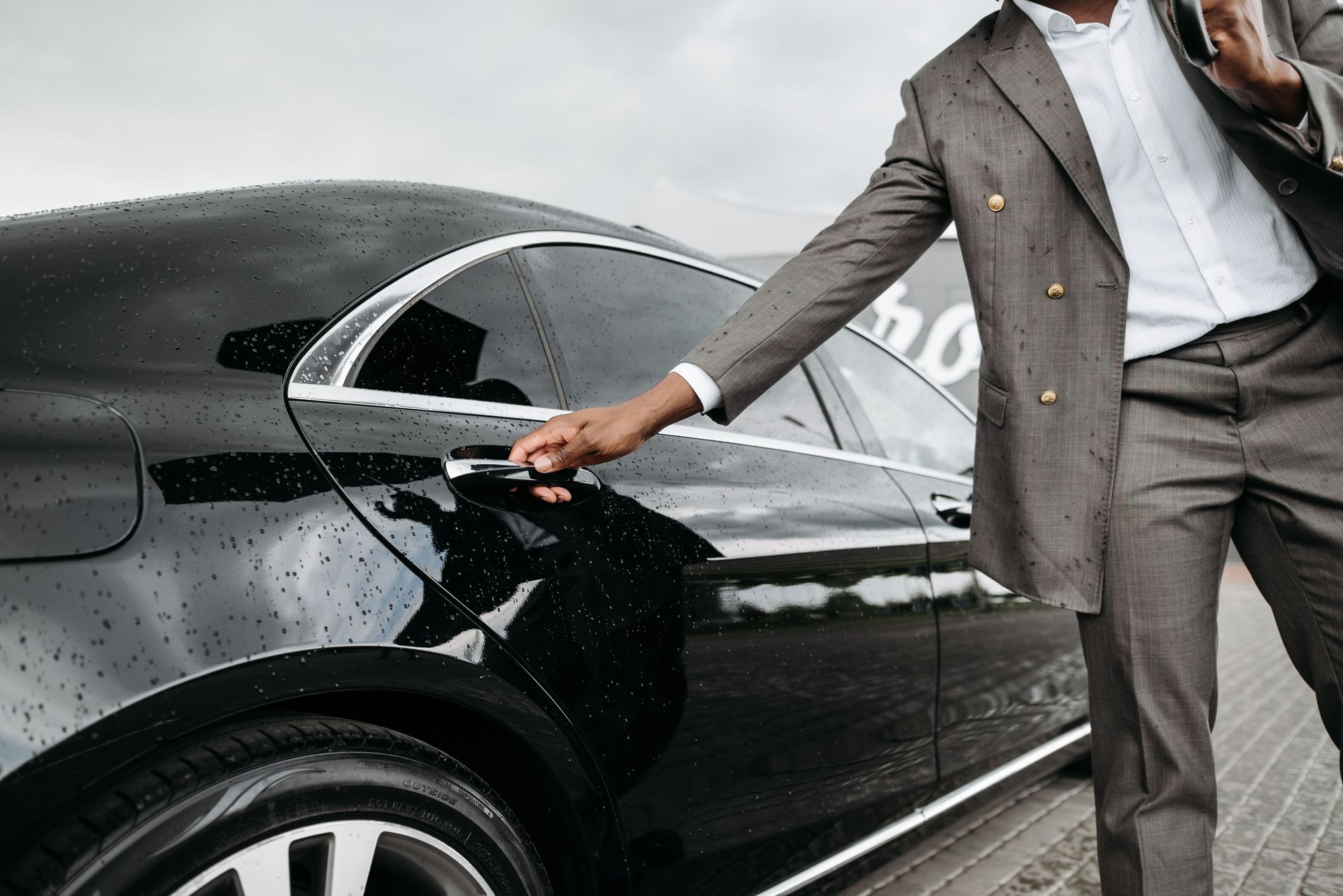 Person in a gray suit opening the door of a black sedan on a rainy day
