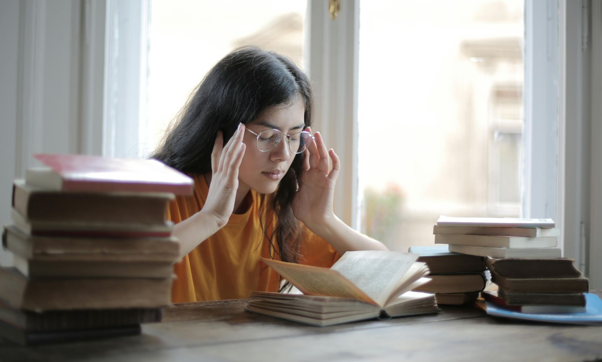 Woman with glasses rubs temples, surrounded by books at a table by a window.