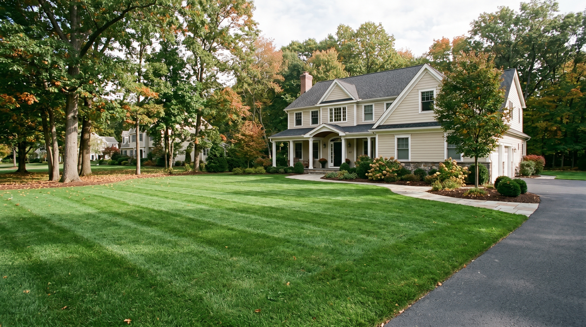 Large gray house with a porch beside a green lawn and driveway, surrounded by trees in autumn.