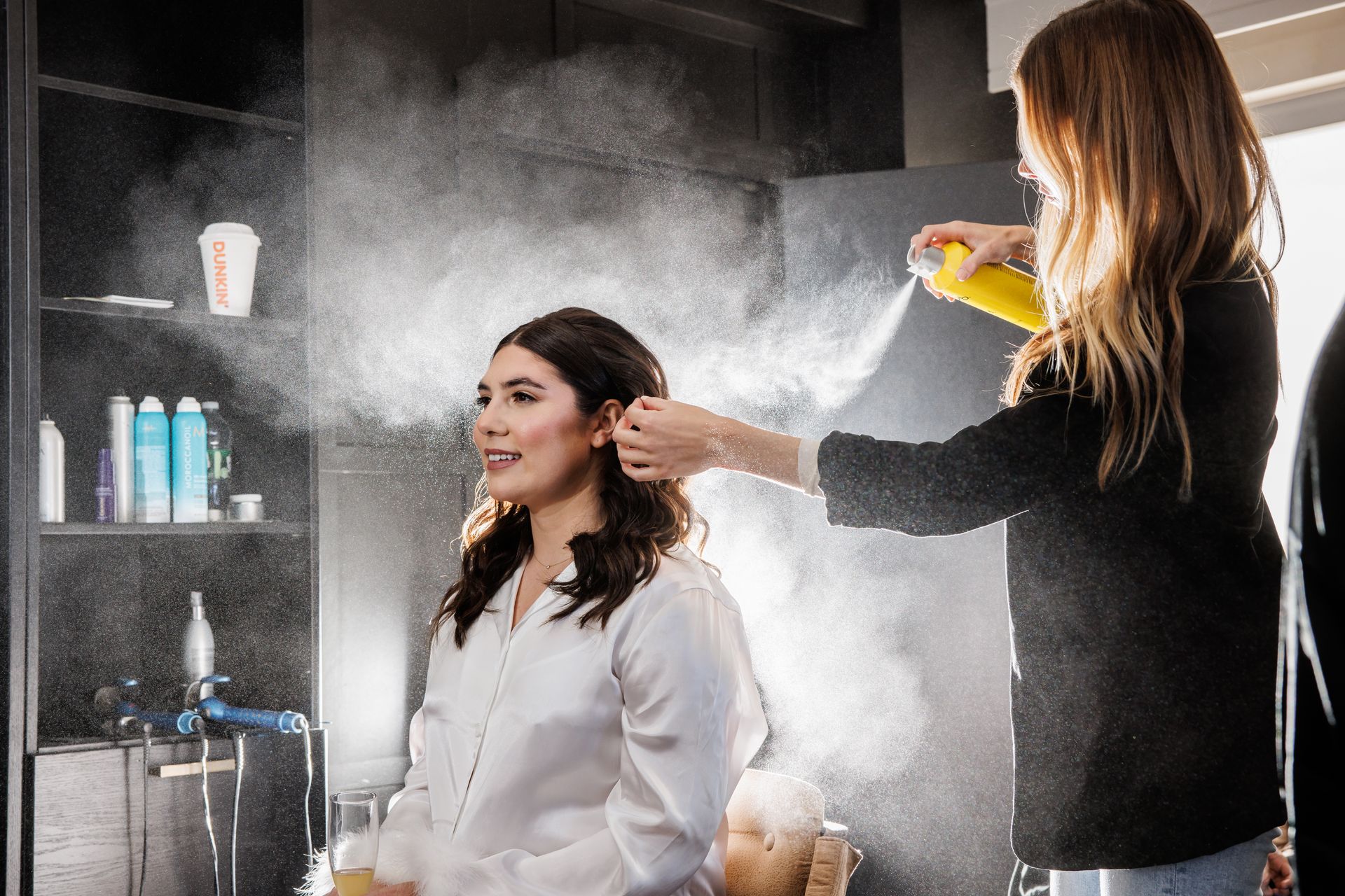Stylist spraying hairspray on seated client in a salon with shelves and smoke-like mist in the background