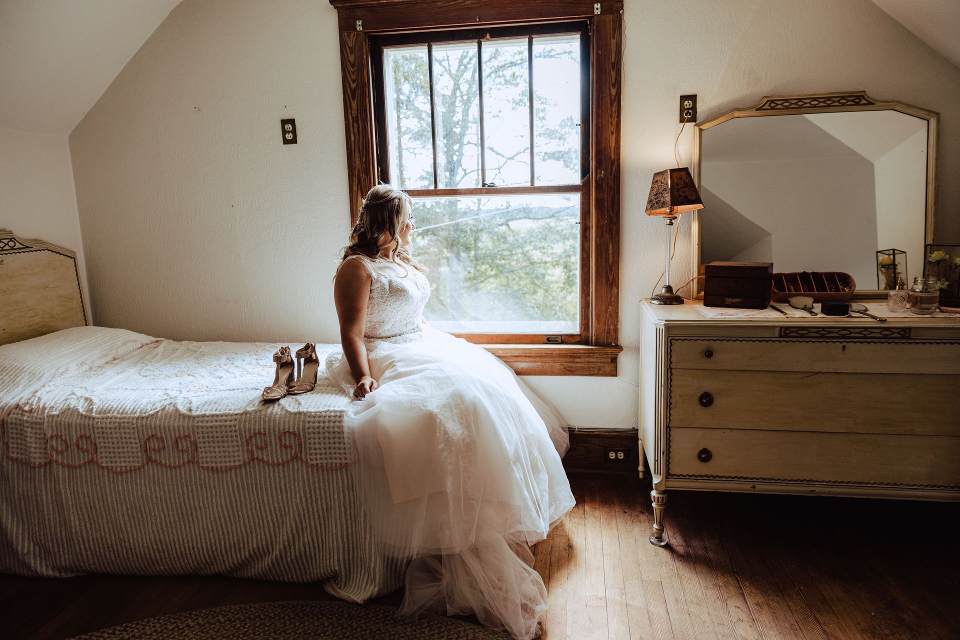 Woman in a white dress seated by a window in a cozy bedroom, with bed and dresser nearby