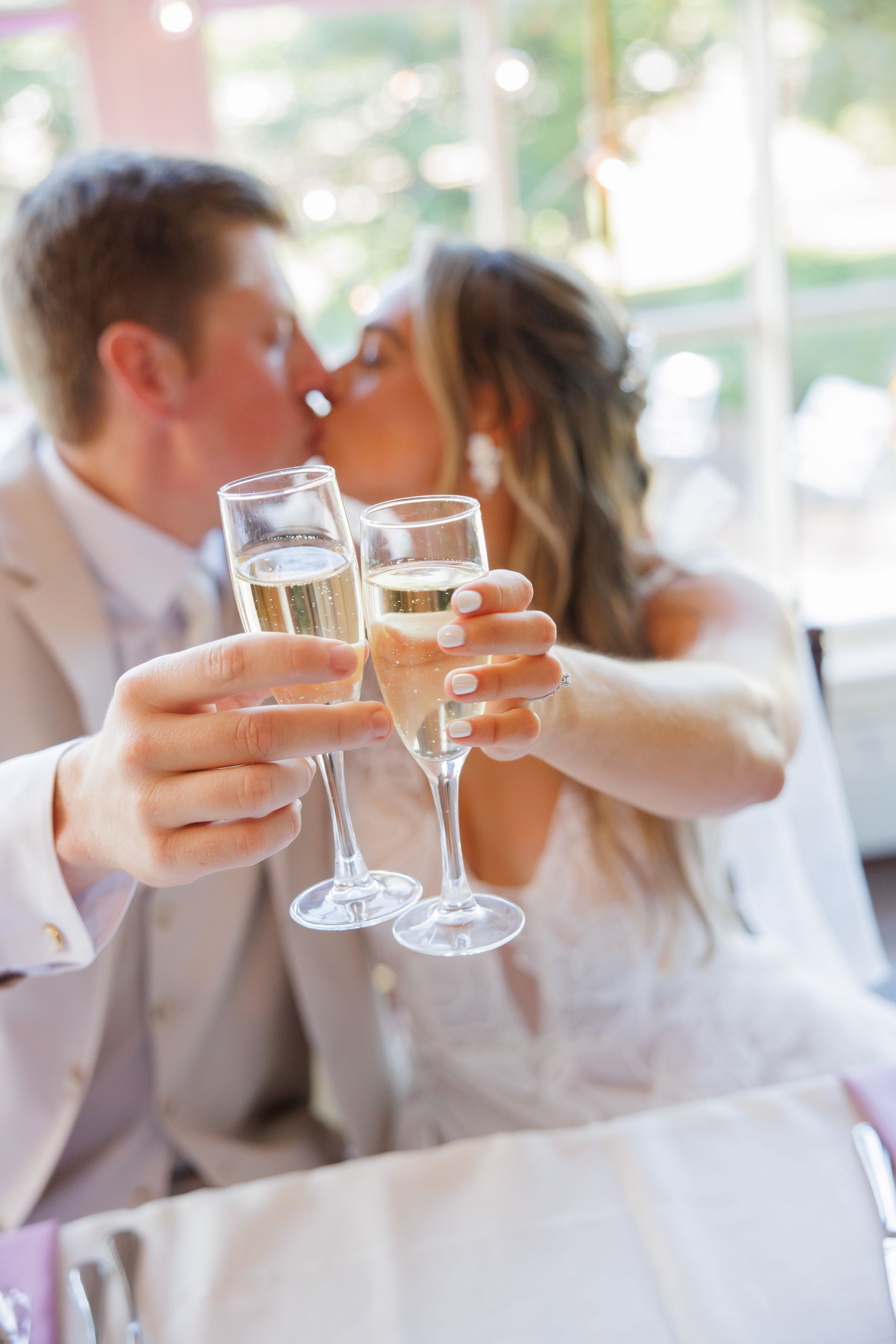 Two people clink champagne glasses while kissing at a wedding reception