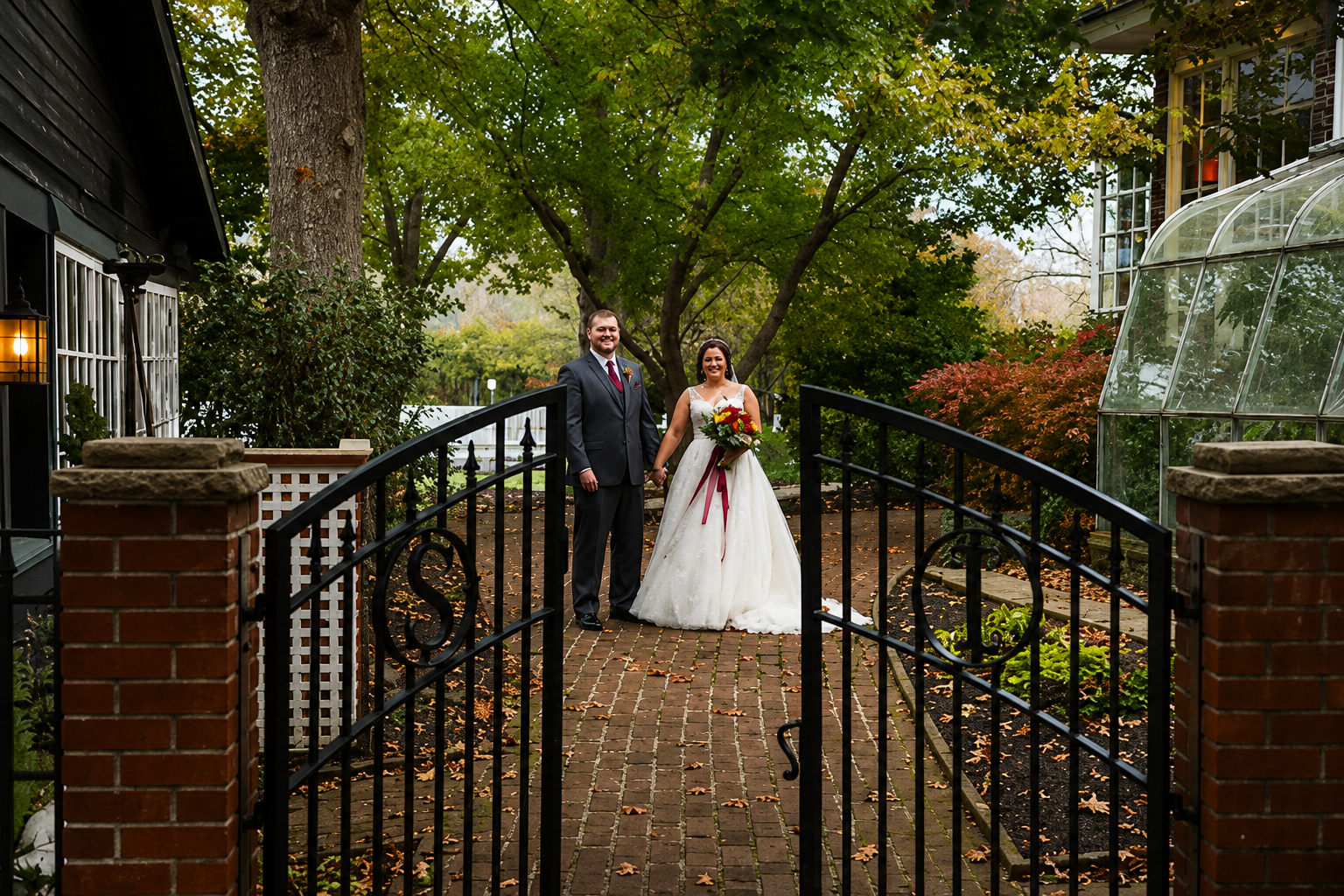Bride and groom standing by a black gate in a garden path with autumn trees and brick walls
