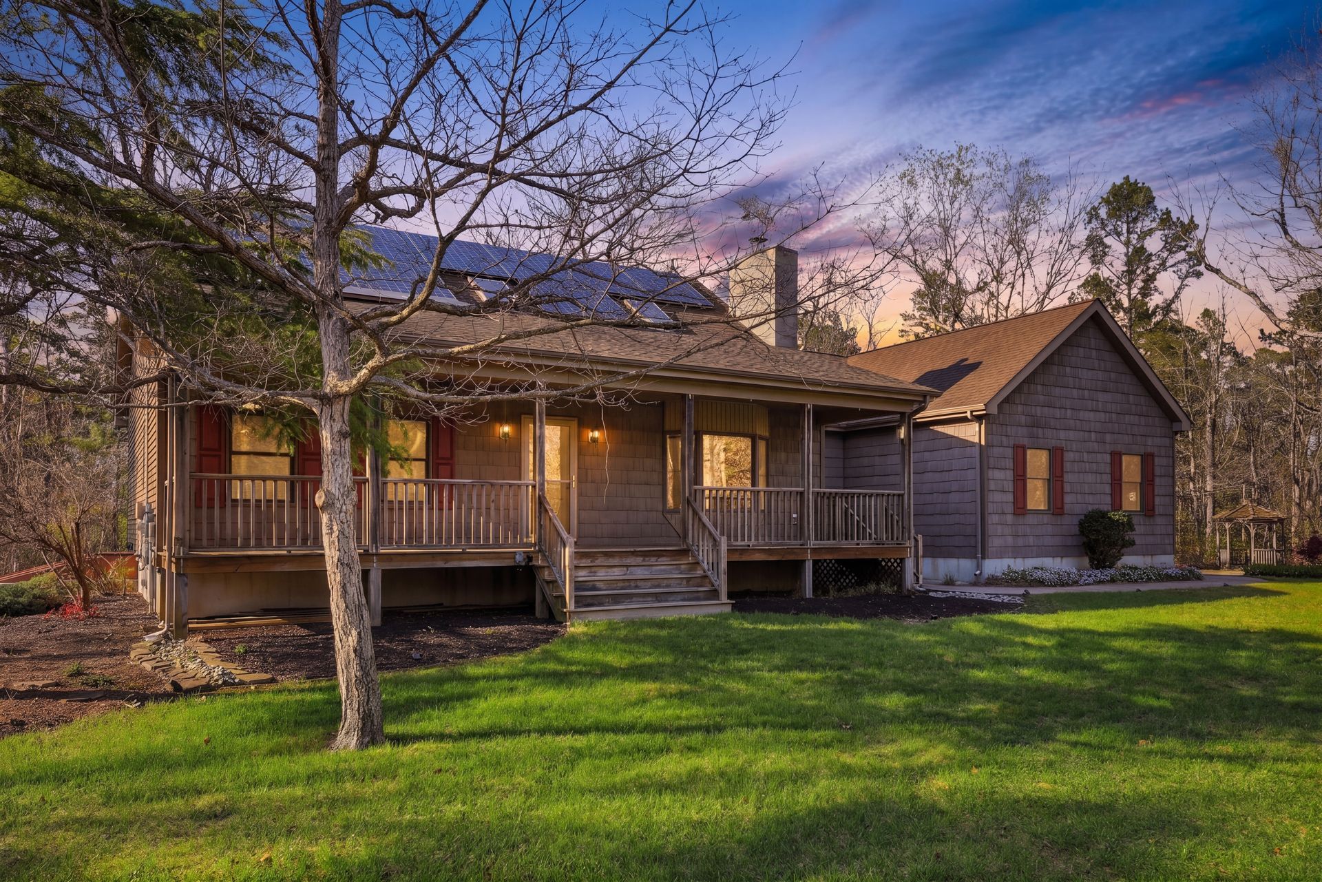 Cozy rustic house with a front porch, lit windows, and a large green lawn at dusk