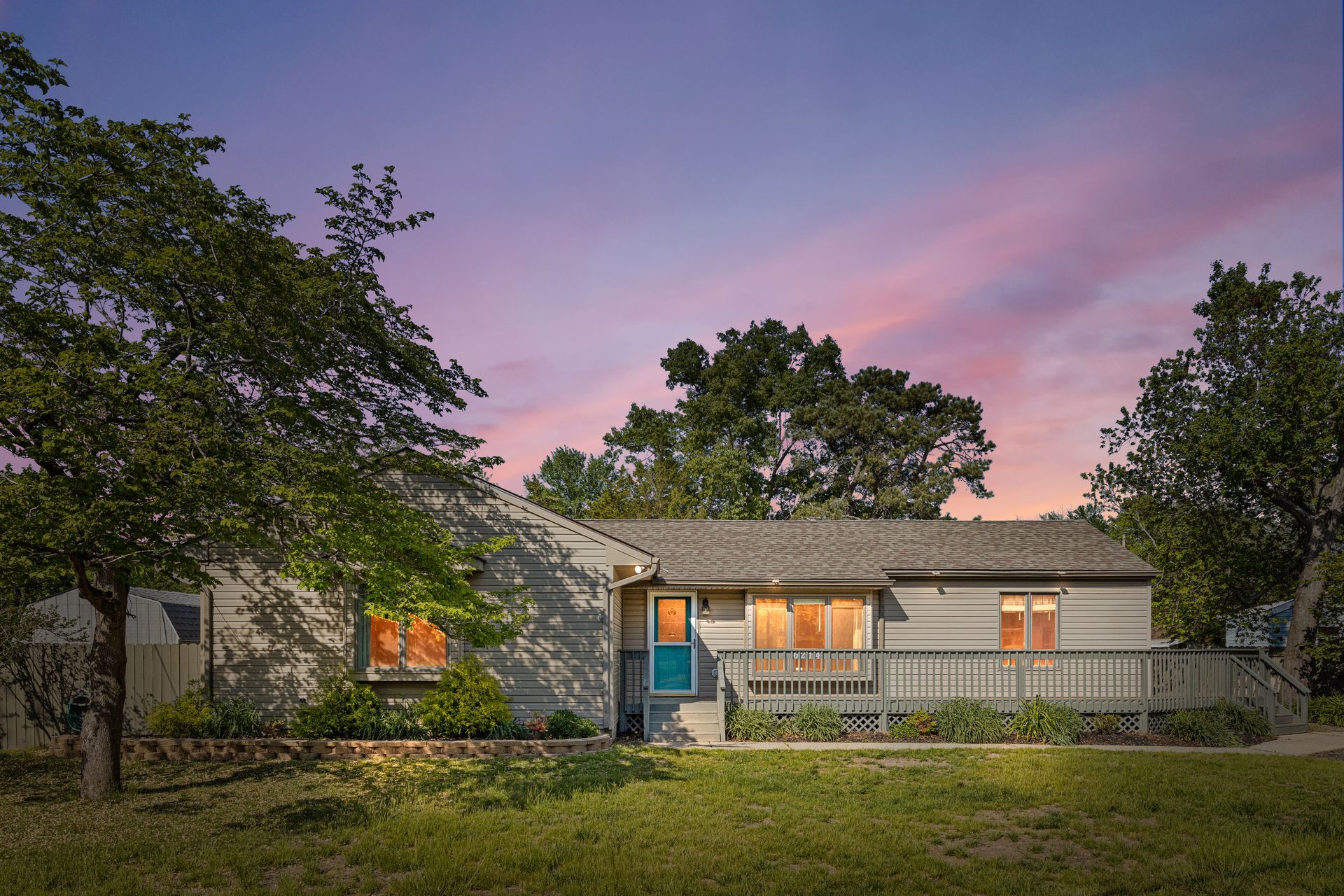 Single-story stone house with lit windows at dusk, framed by trees and a pink-purple sky.