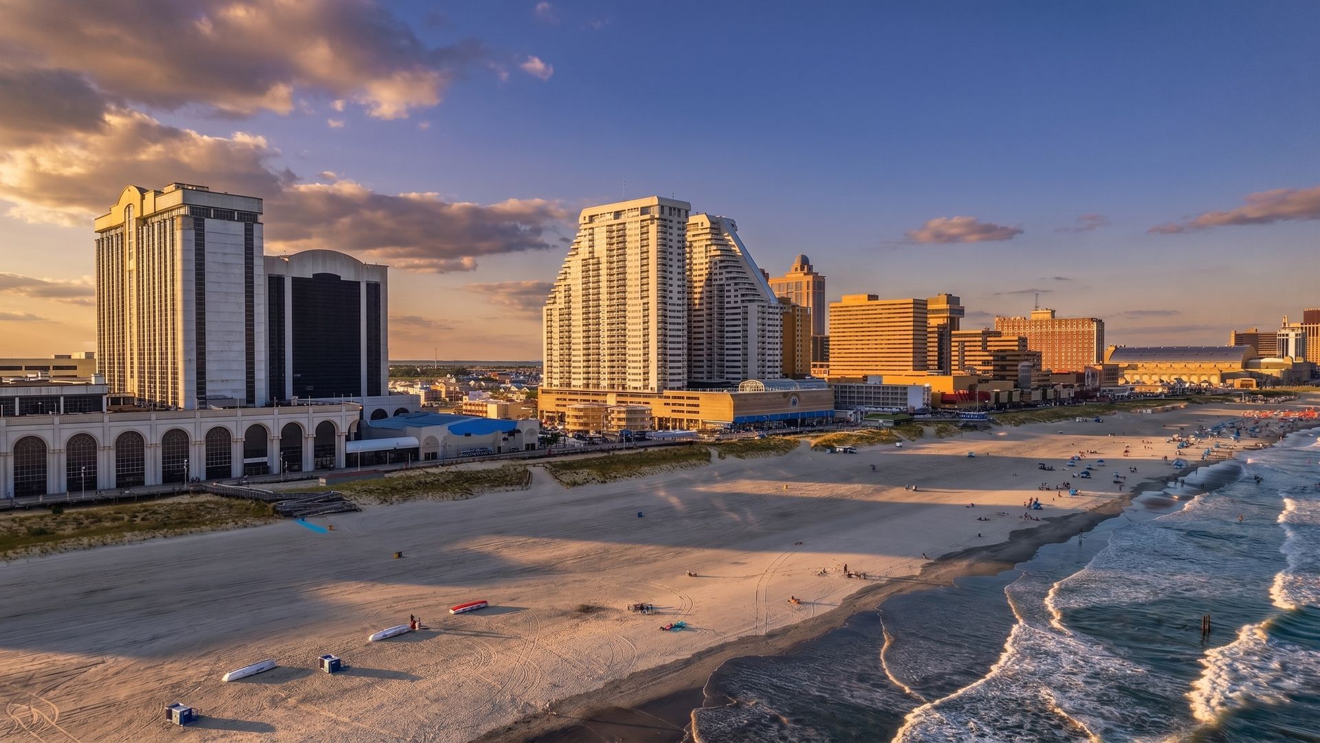 Coastal city skyline at sunset with high-rise buildings beside a sandy beach and waves.