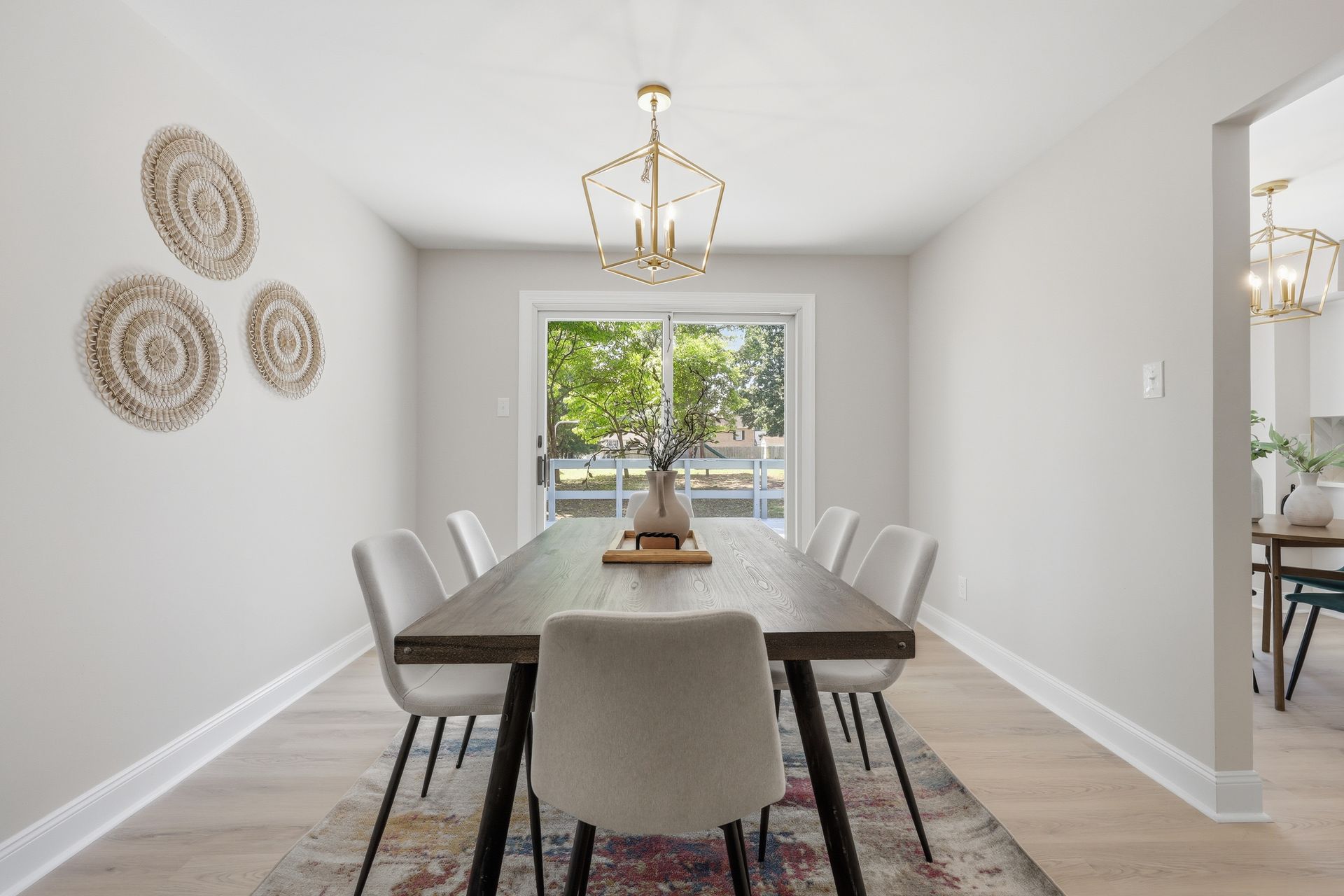 Bright dining room with a long table, gray chairs, chandelier, and sliding glass door to a backyard.
