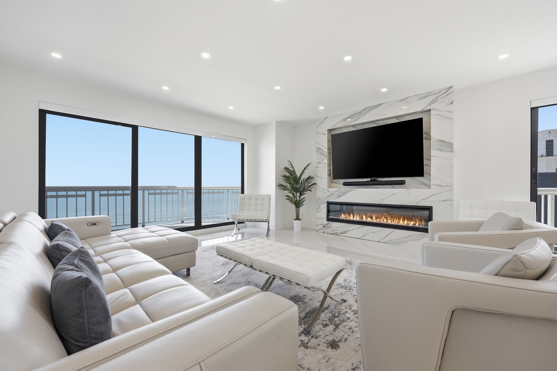 Bright modern living room with white sofas, fireplace, wall-mounted TV, and large ocean-view windows