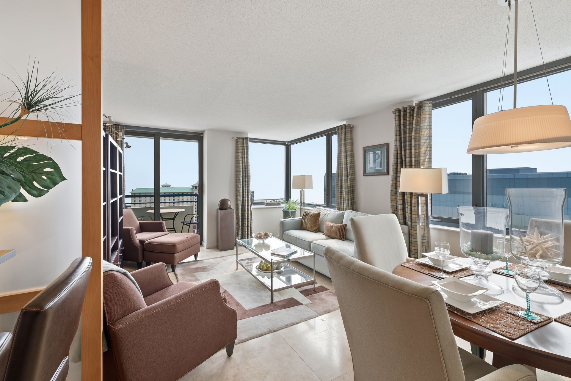 Bright living-dining room with city-view windows, brown chairs, white sofa, and glass dining table