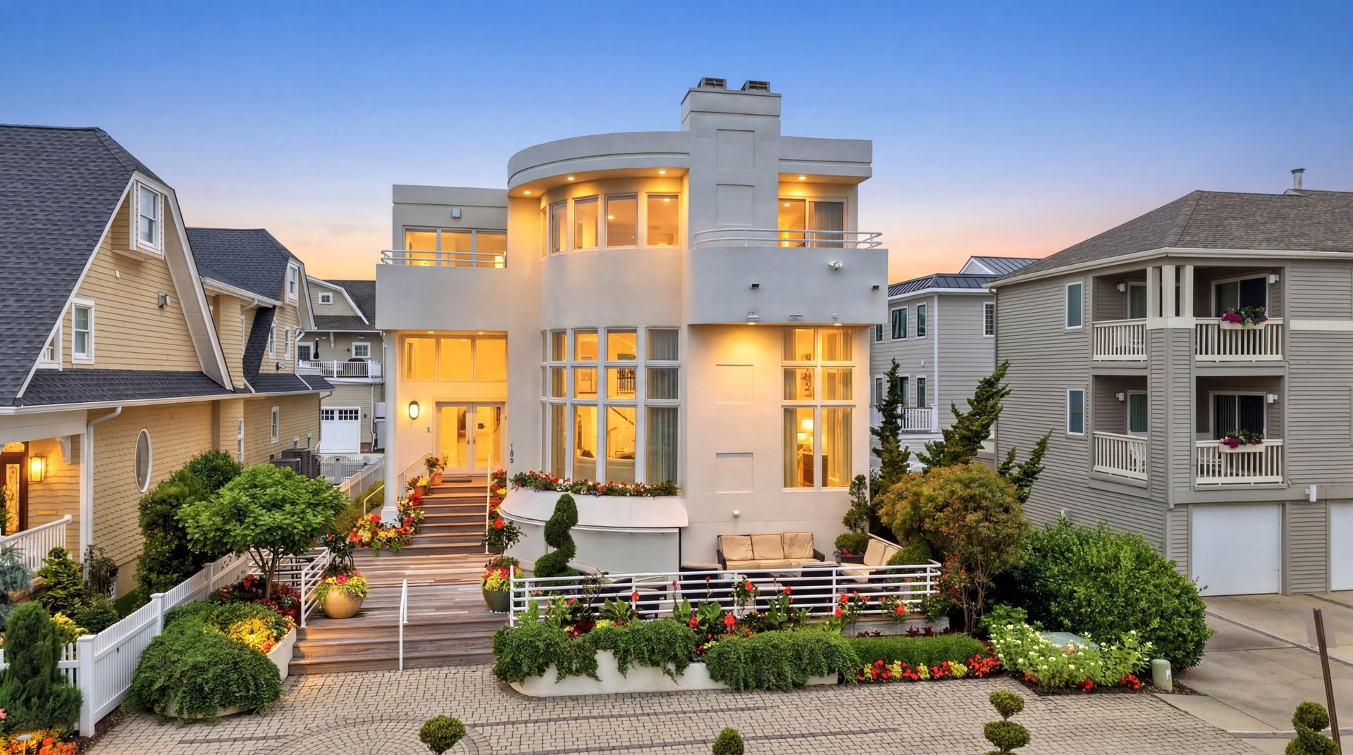 Modern white two-story house with warm lights at dusk, front garden, and neighboring homes on both sides