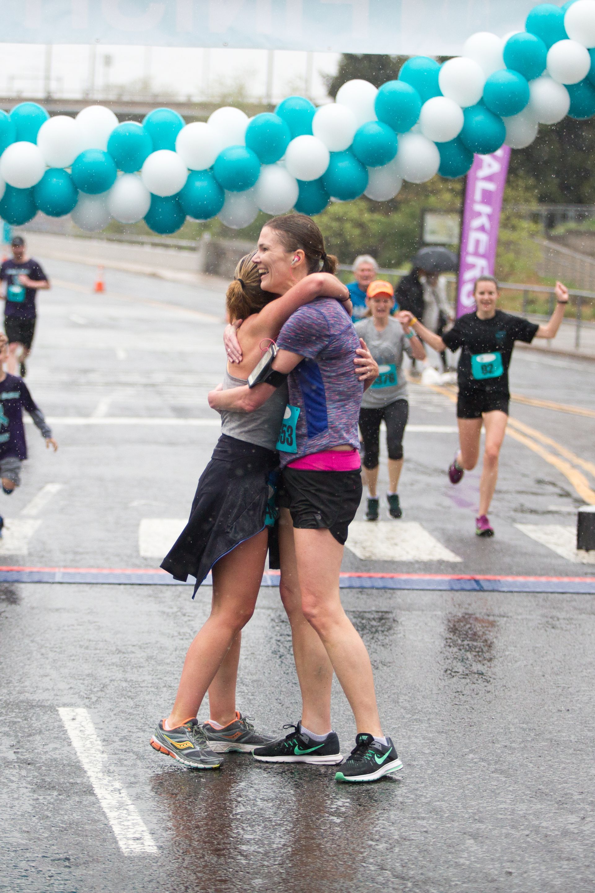 Two runners hugging at a rainy finish line beneath blue and white balloons