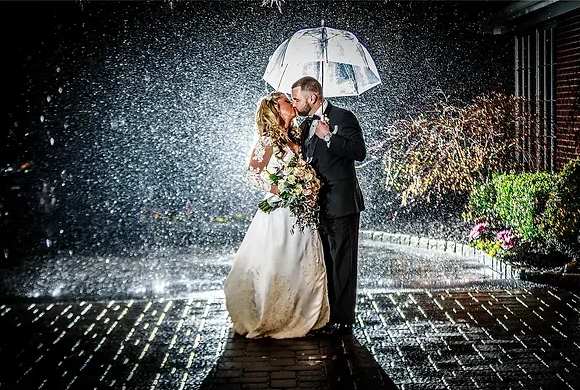 Couple kissing under an umbrella in heavy rain, standing on a wet cobblestone path at night.