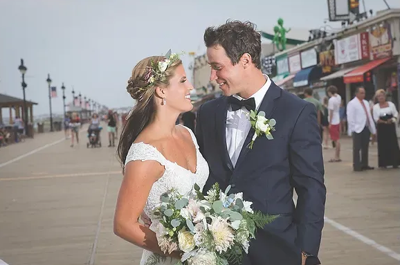 Newlyweds smiling on a seaside boardwalk, holding a bouquet and wearing formal wedding attire