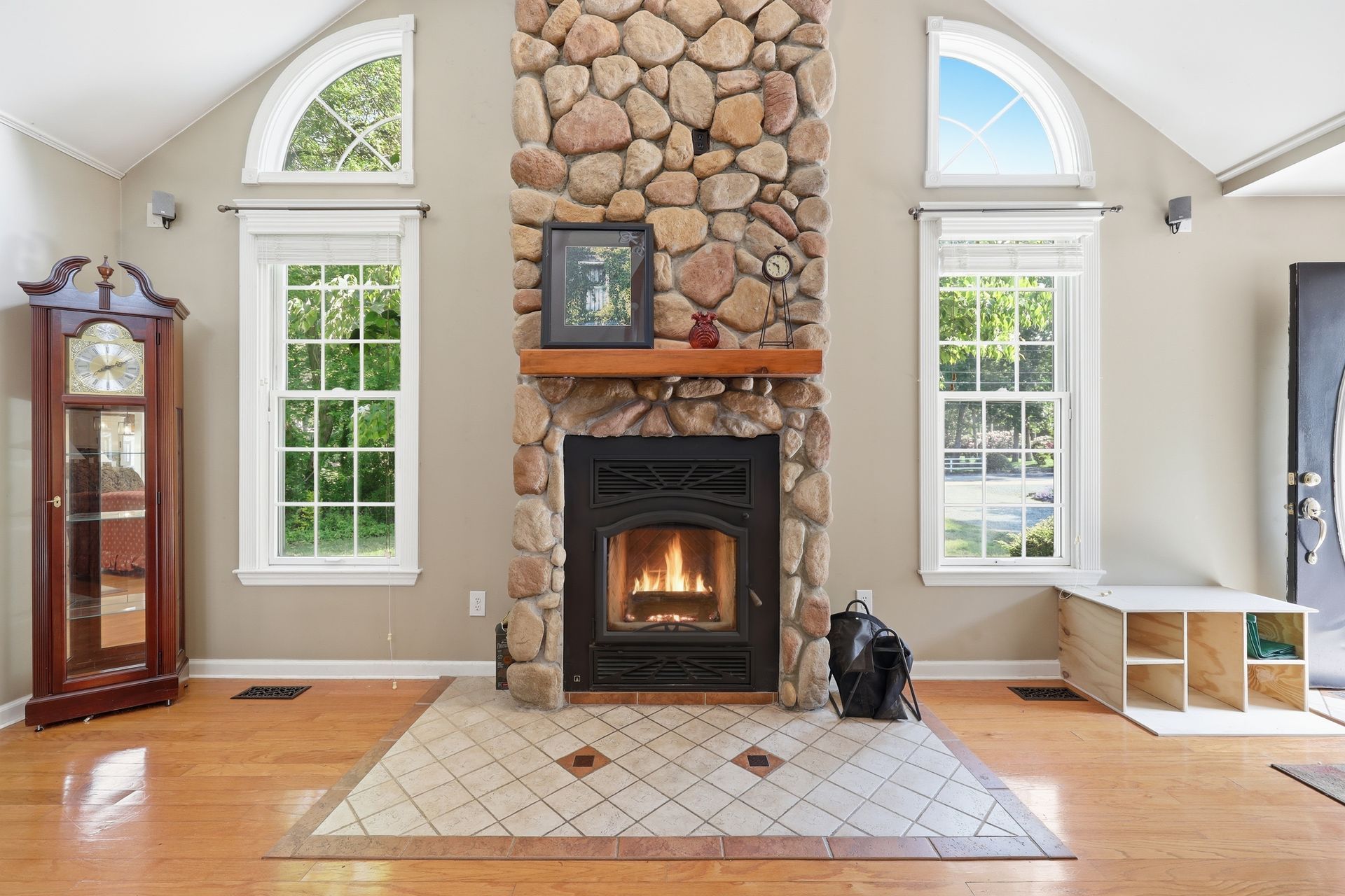 Living room with stone fireplace, lit fire, tall windows, and hardwood floors