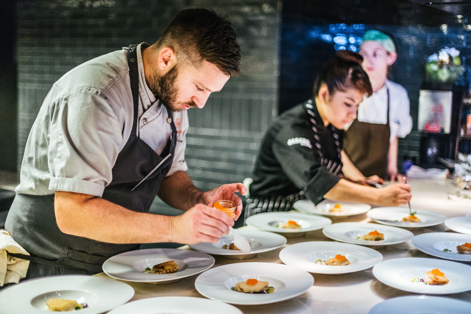 Chef plating dishes in a commercial kitchen with several white plates arranged on a counter