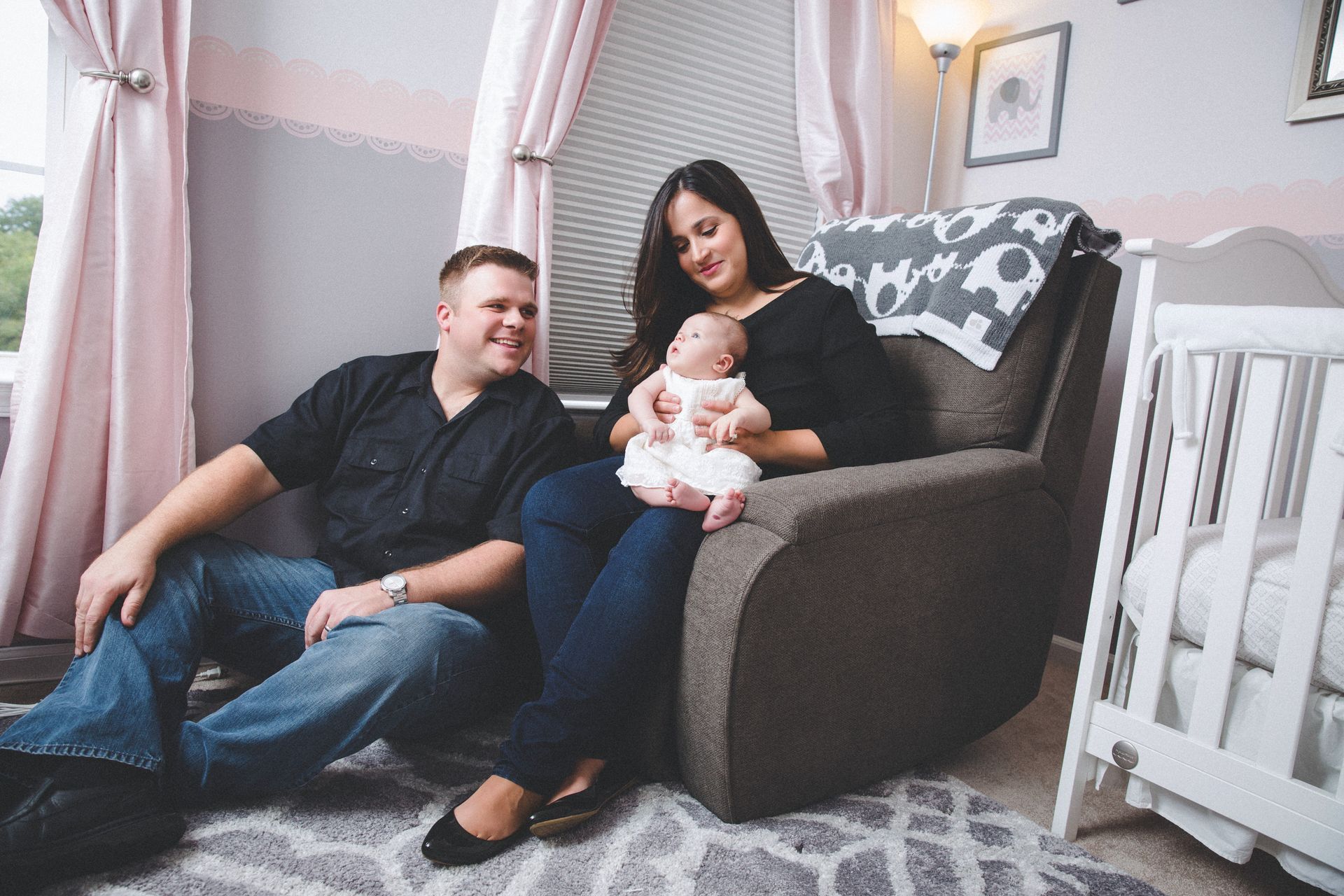 Parents with baby in a cozy nursery, sitting by a white crib and patterned armchair.