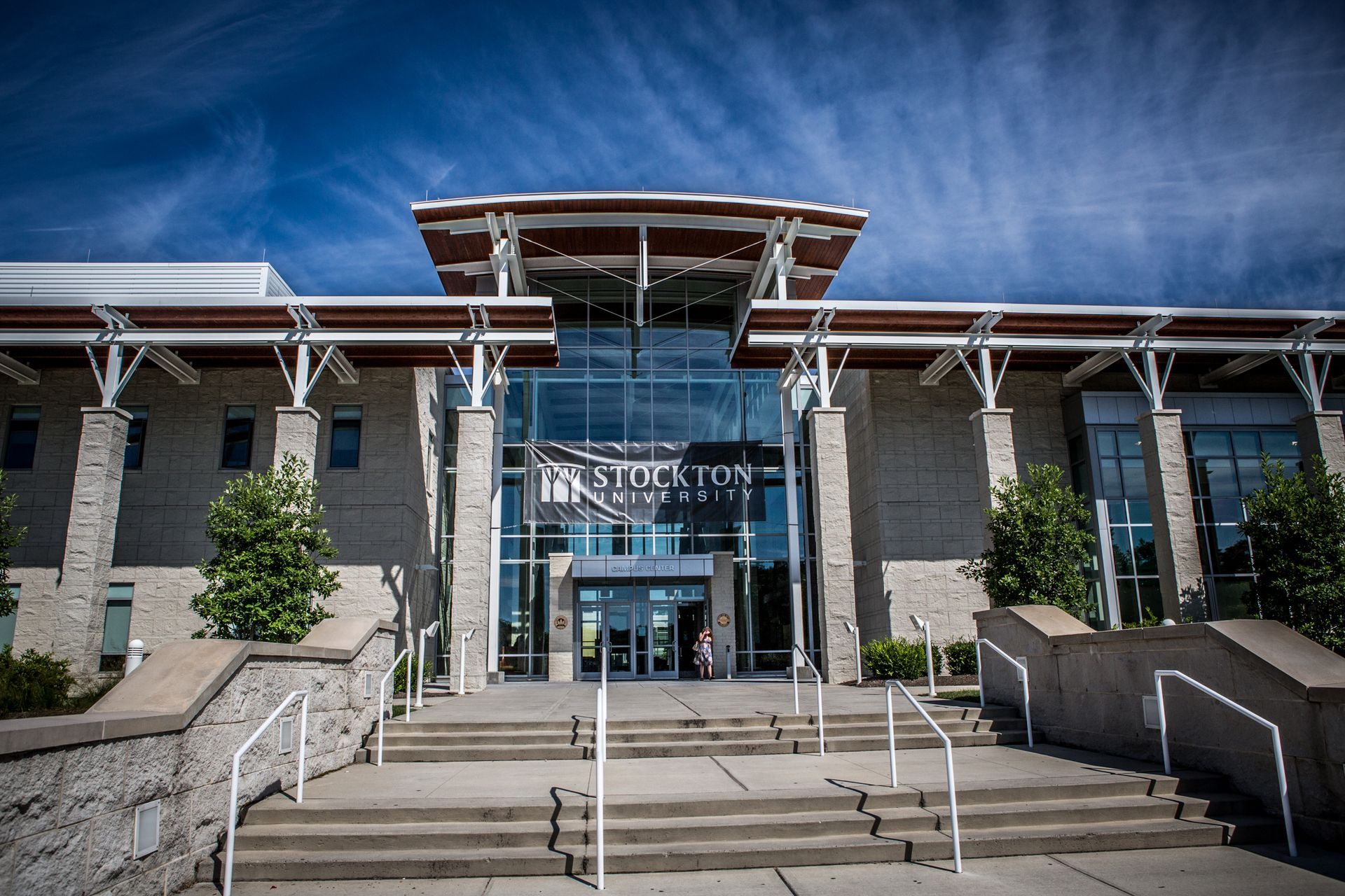 Modern stone building entrance with glass doors, steps, railings, and blue sky above