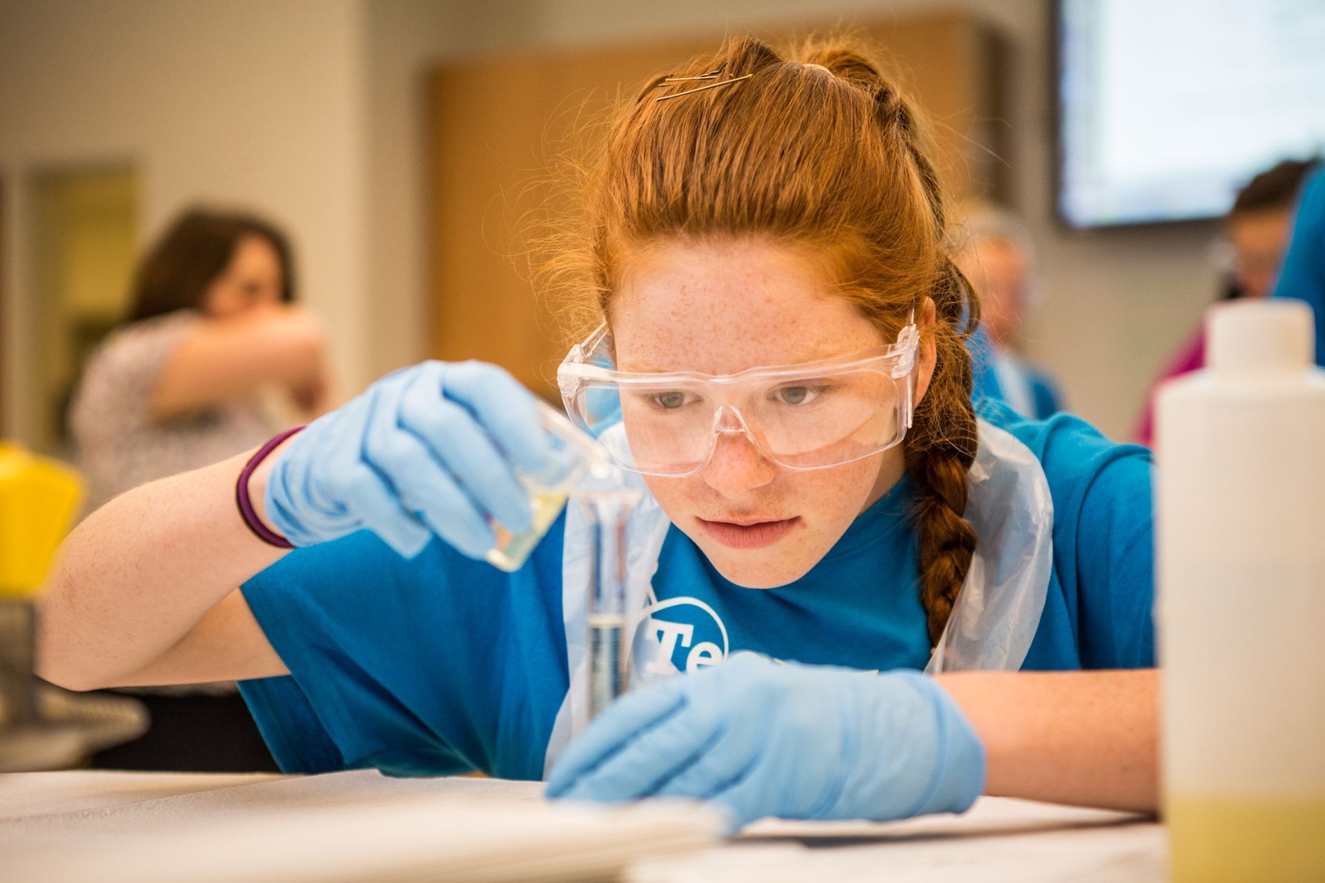 Gloved lab worker in safety goggles handling a test tube at a table in a science lab