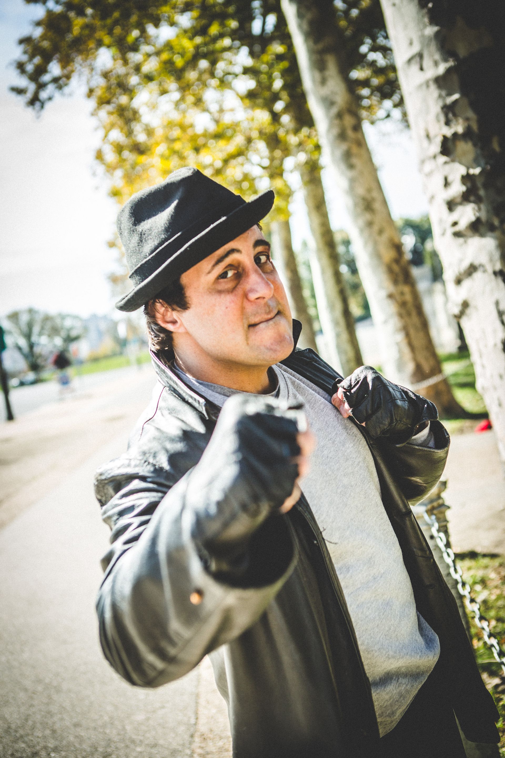 Man in a black hat and coat posing outdoors, reaching toward the camera near trees