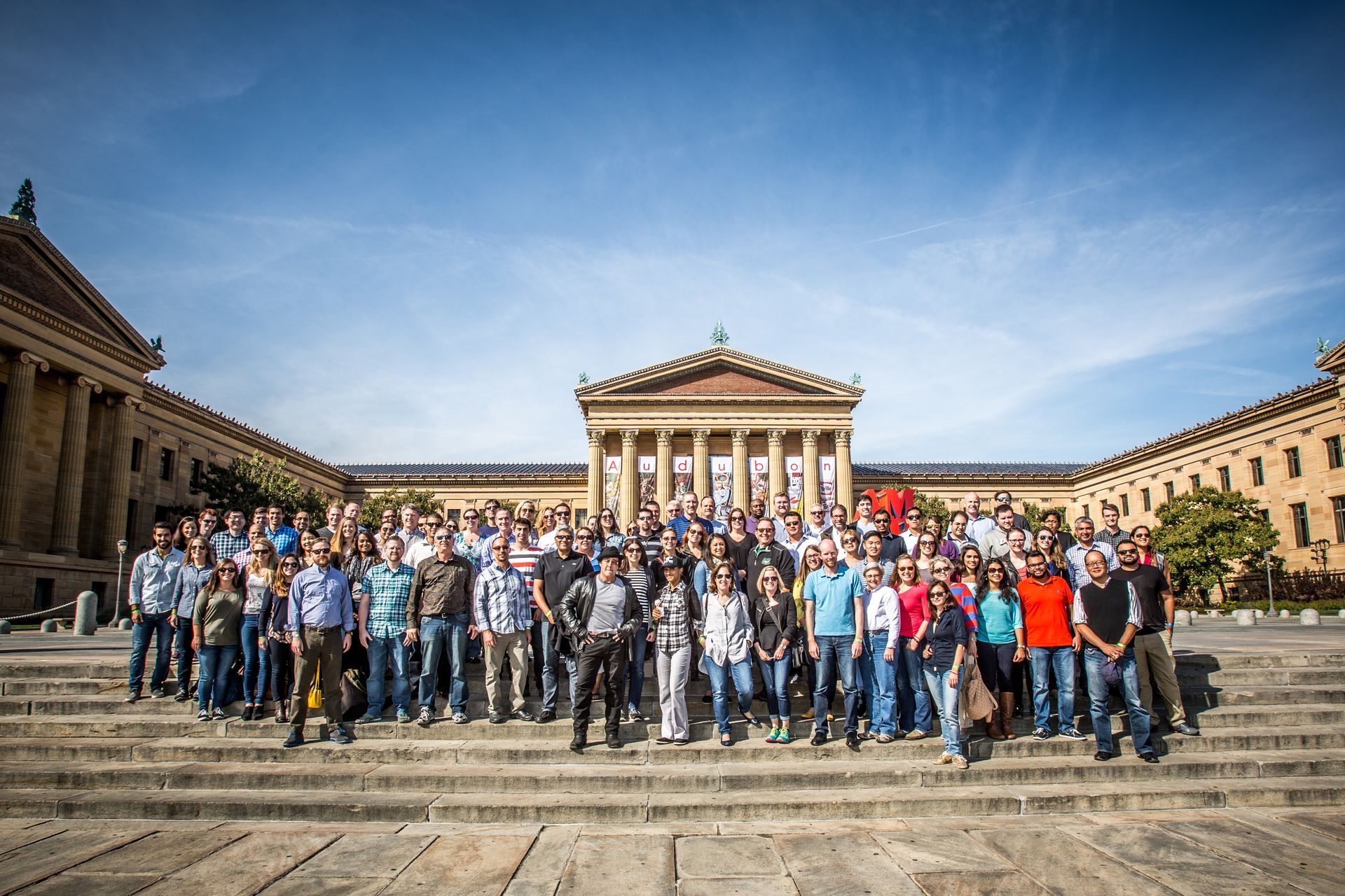 Large group posing in front of a classical building with columns under a bright blue sky