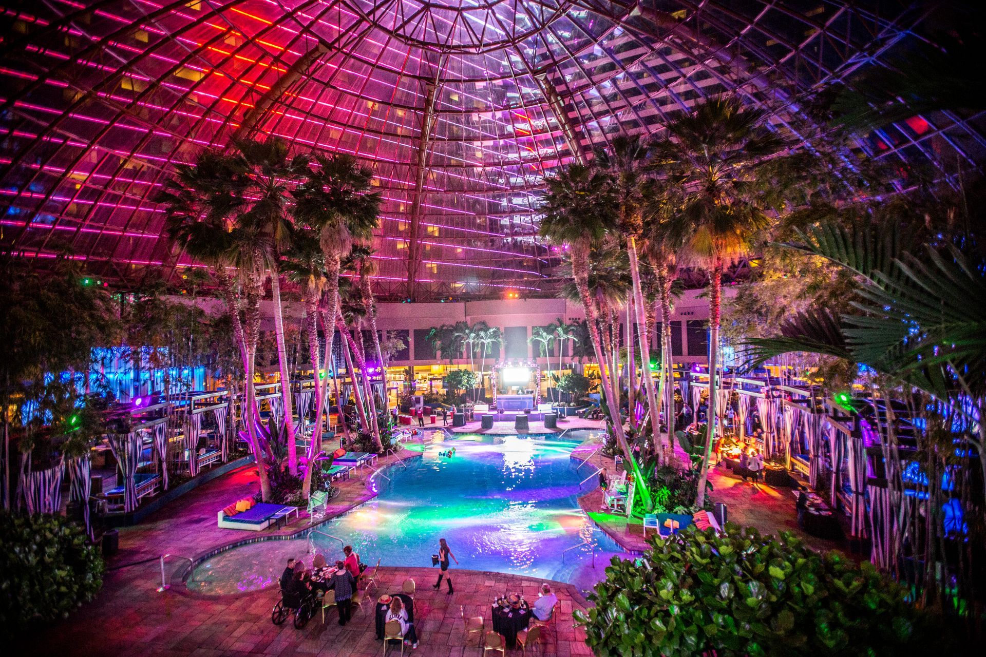 Colorful indoor atrium with a glowing pool, palm trees, and a glass dome ceiling at night