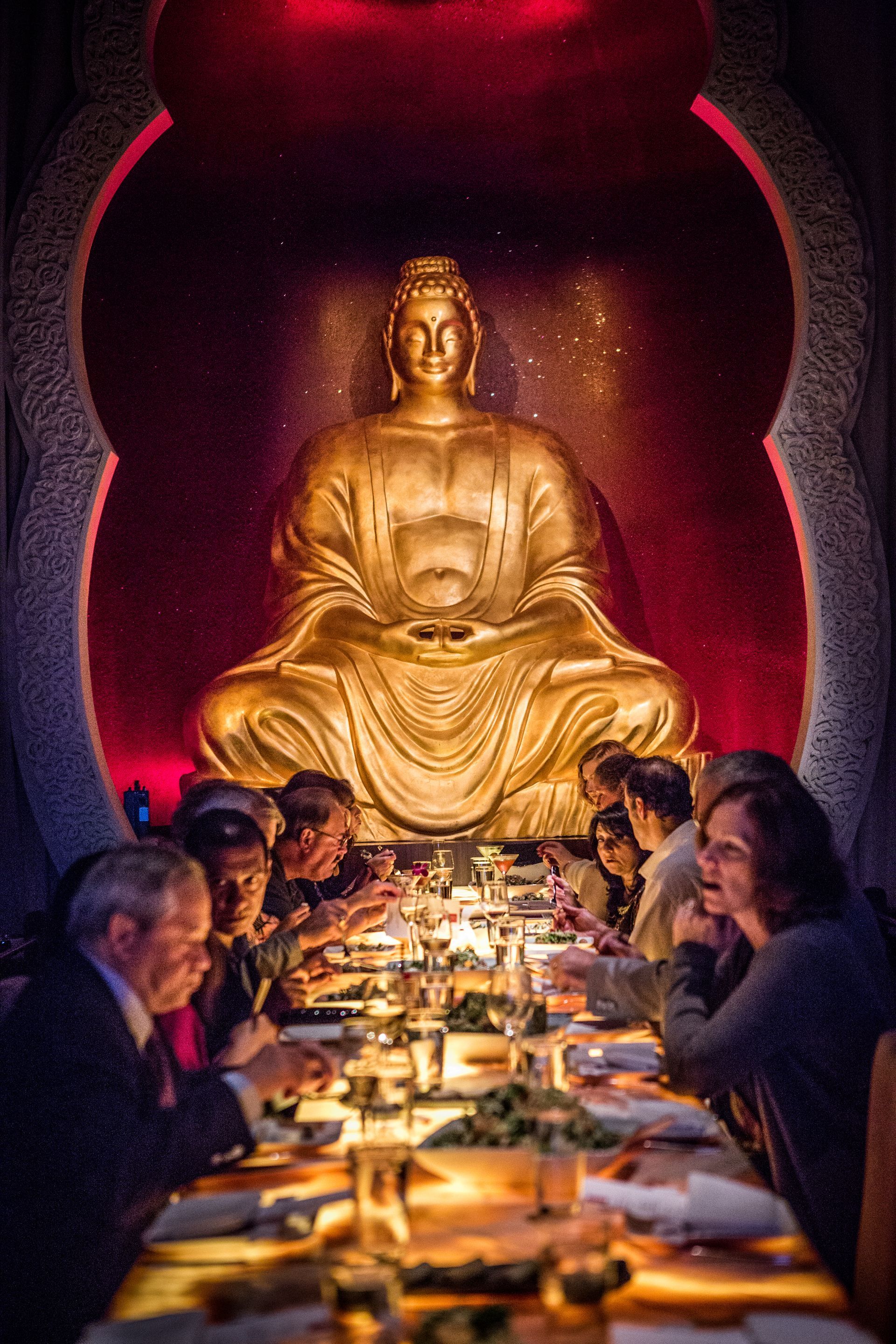 Dining table with people eating beneath a large golden Buddha statue in a dim, red-lit room