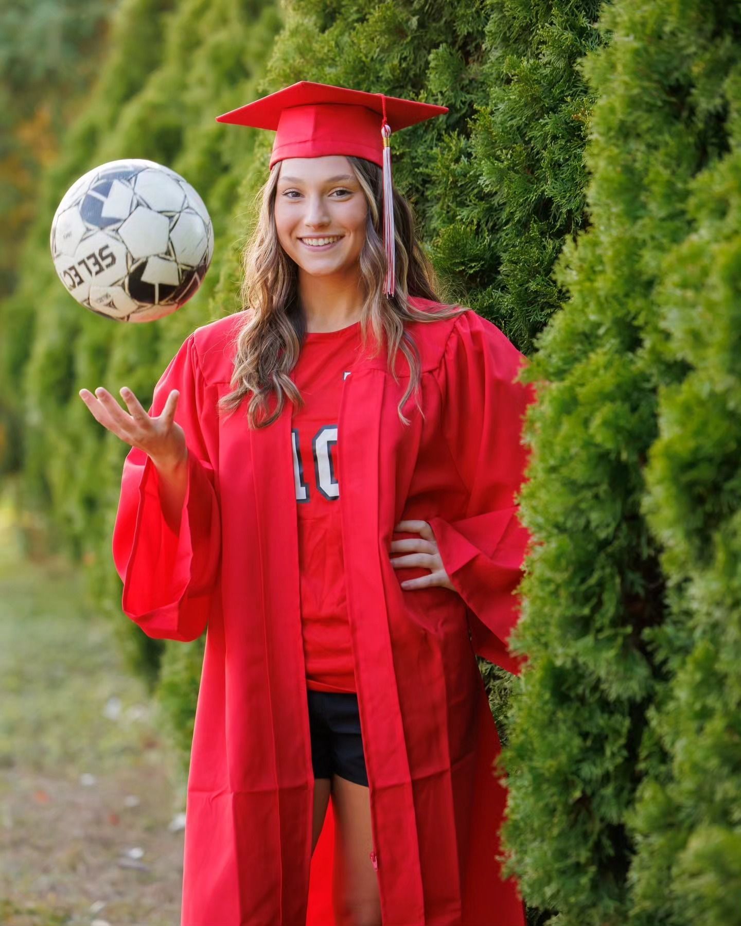 Person in red graduation cap and gown, smiling outdoors and balancing a soccer ball on one hand.