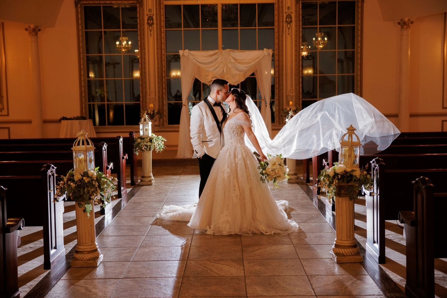 Bride and groom pose in a warmly lit church aisle, with a flowing veil and floral decorations.