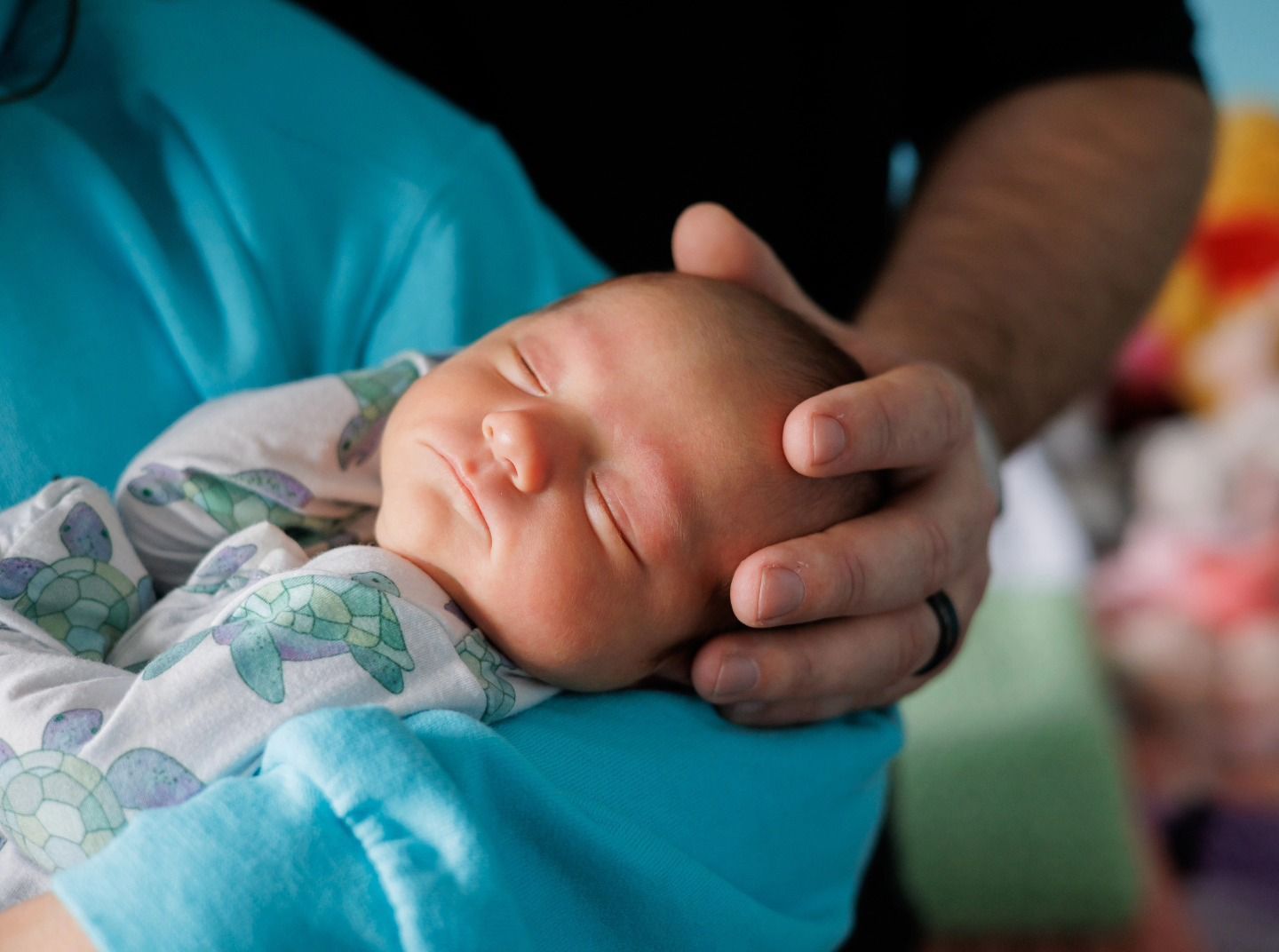 Sleeping newborn wrapped in a teal blanket, resting in an adult’s hand and arms