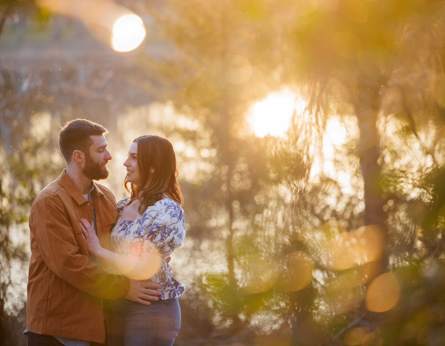 Couple standing close in a sunlit forest, smiling at each other with warm golden bokeh in the background.