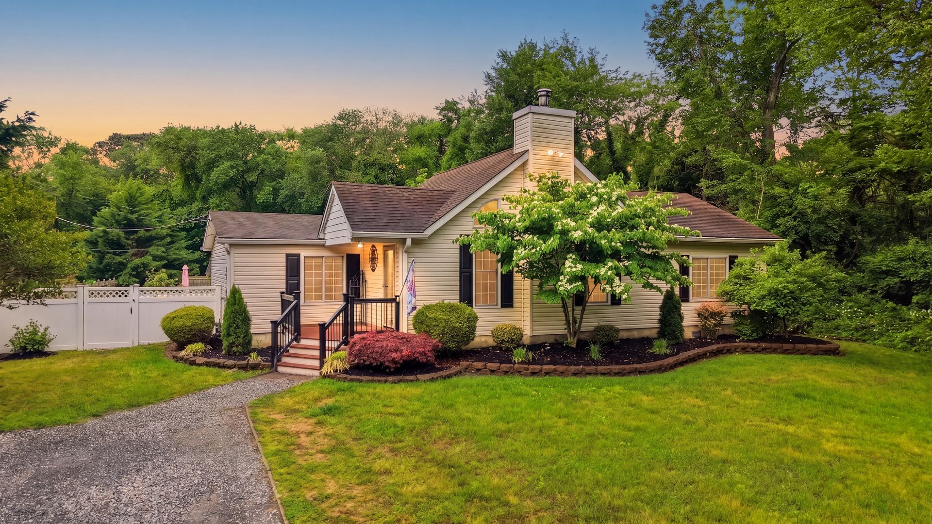 Cozy cottage with lit porch at dusk, surrounded by green lawn and trees.