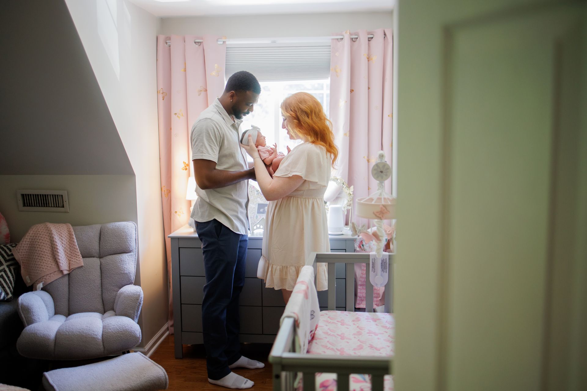 Two adults standing by a crib in a softly lit nursery, looking at a small object together