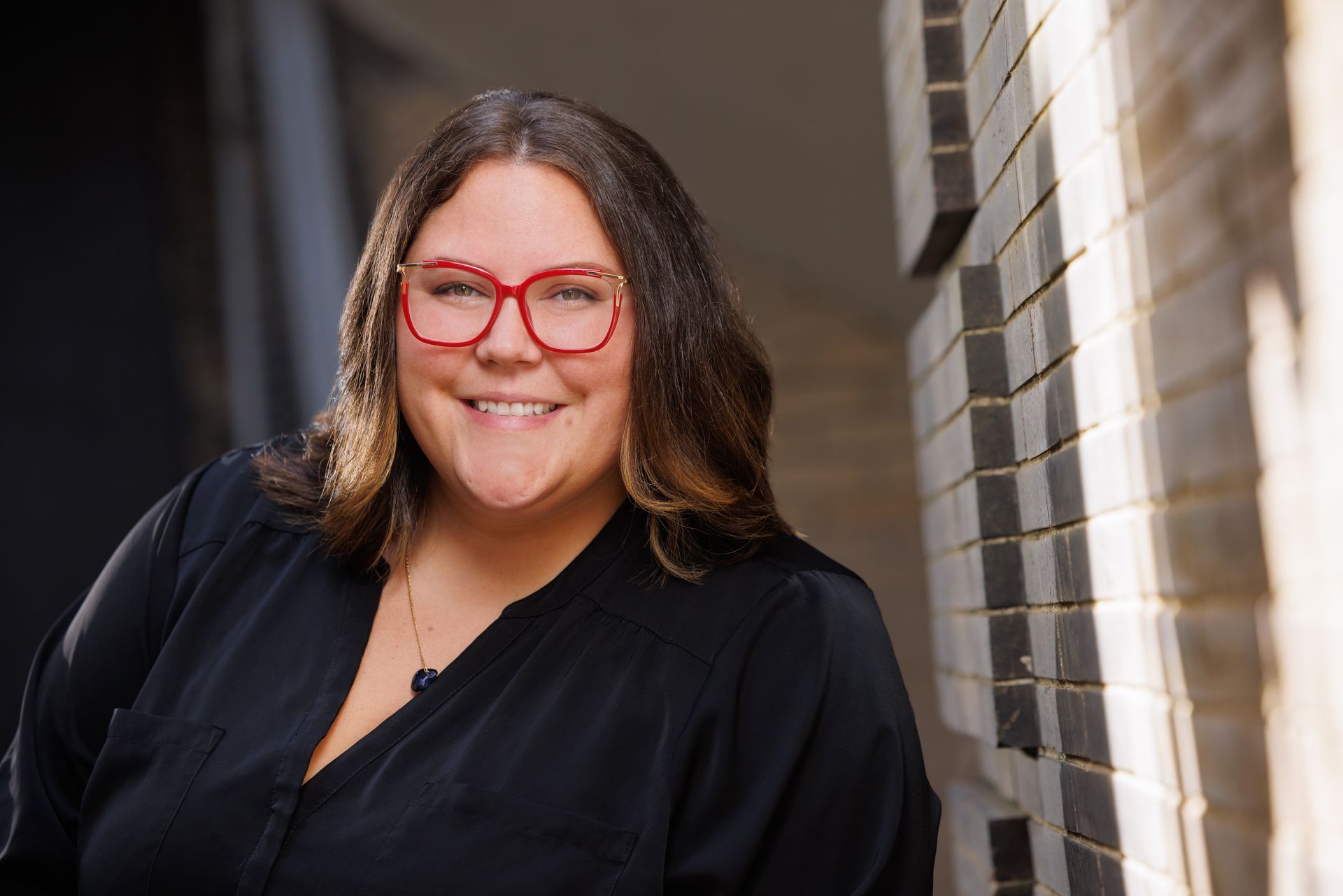 Smiling person with red glasses and dark hair, wearing a black top beside a brick wall outdoors