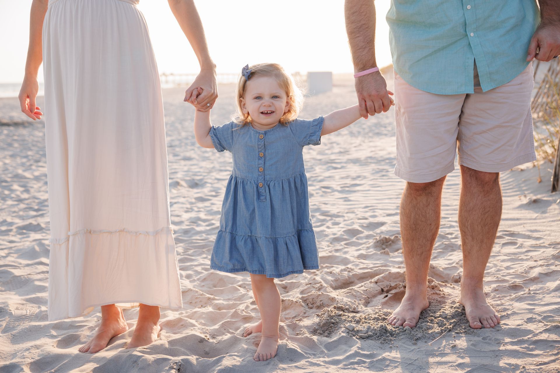Family holding hands on a sandy beach, with a toddler in a blue dress walking between two adults
