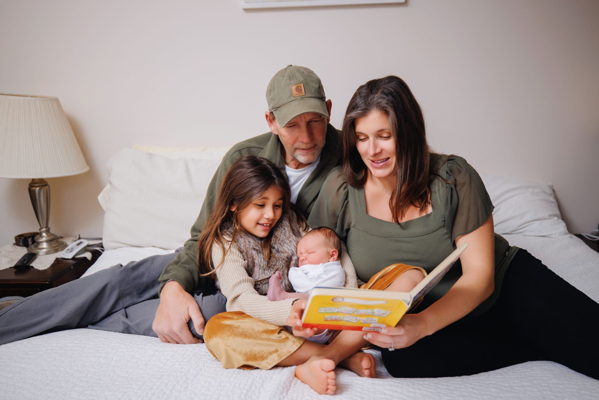 Family sitting on a bed, reading a book together with a newborn baby between them.