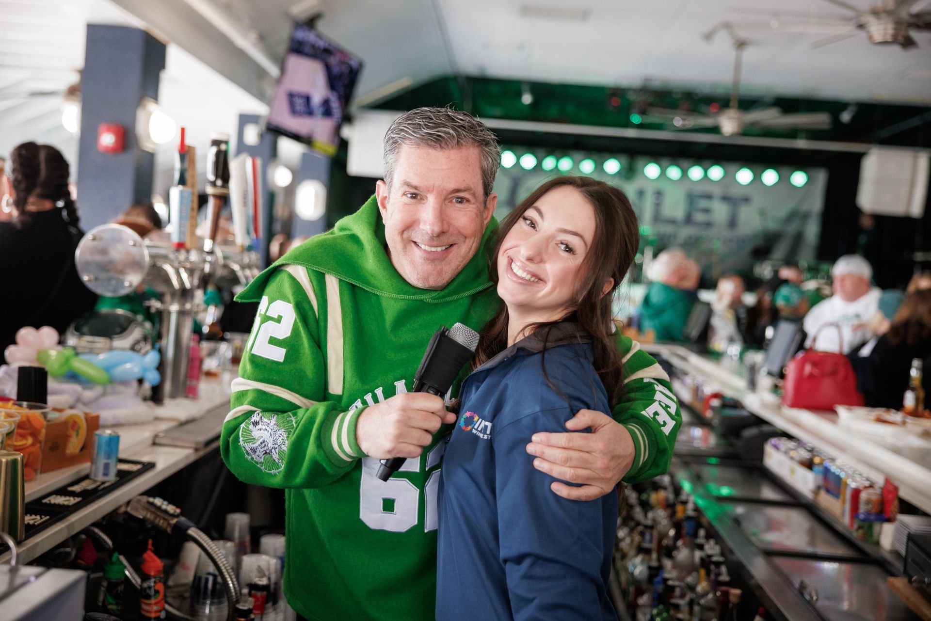 Two smiling people in green and blue jackets posing together at a busy indoor event table