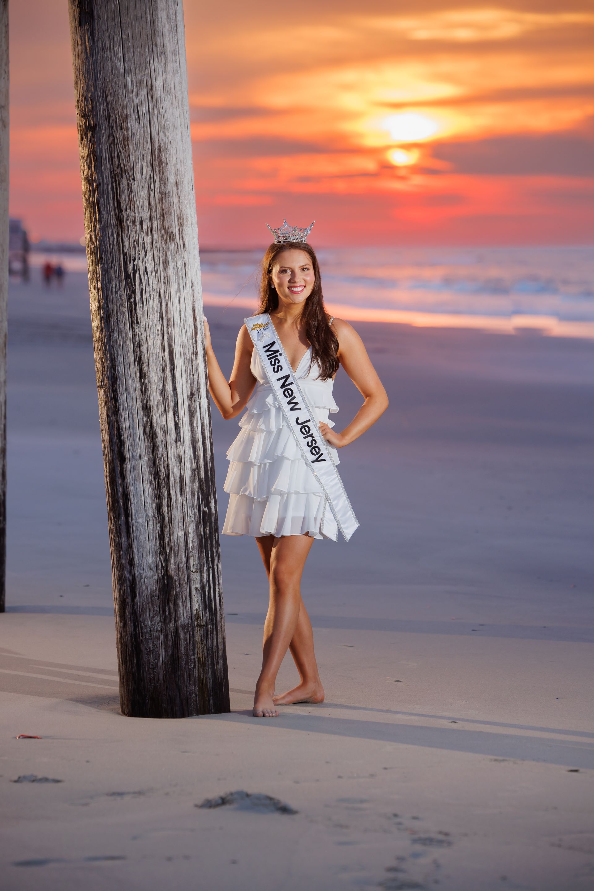 Woman in a white dress standing by a palm tree on a beach at sunset.