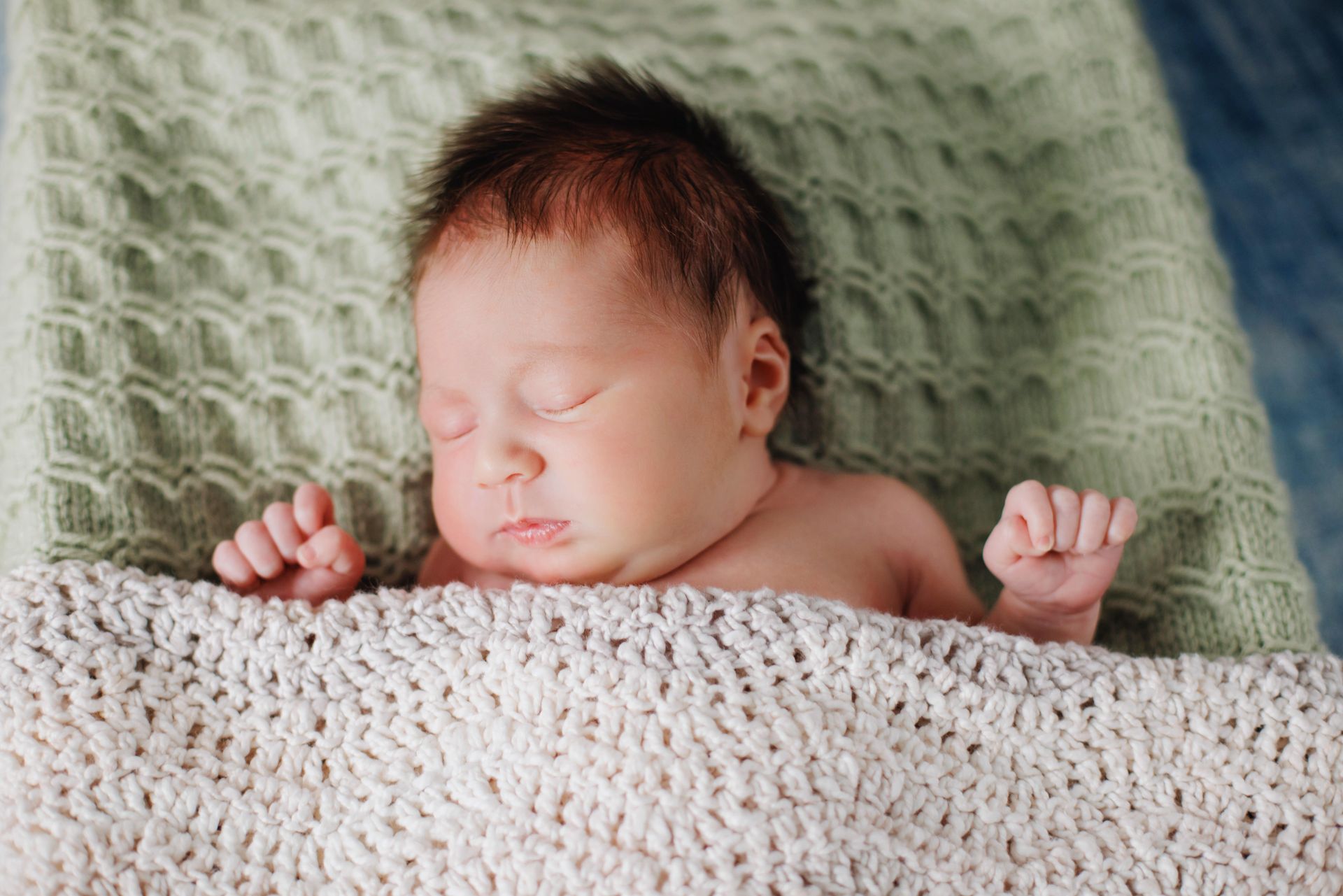 Sleeping newborn wrapped in a white blanket on a green knitted blanket, with tiny hands visible