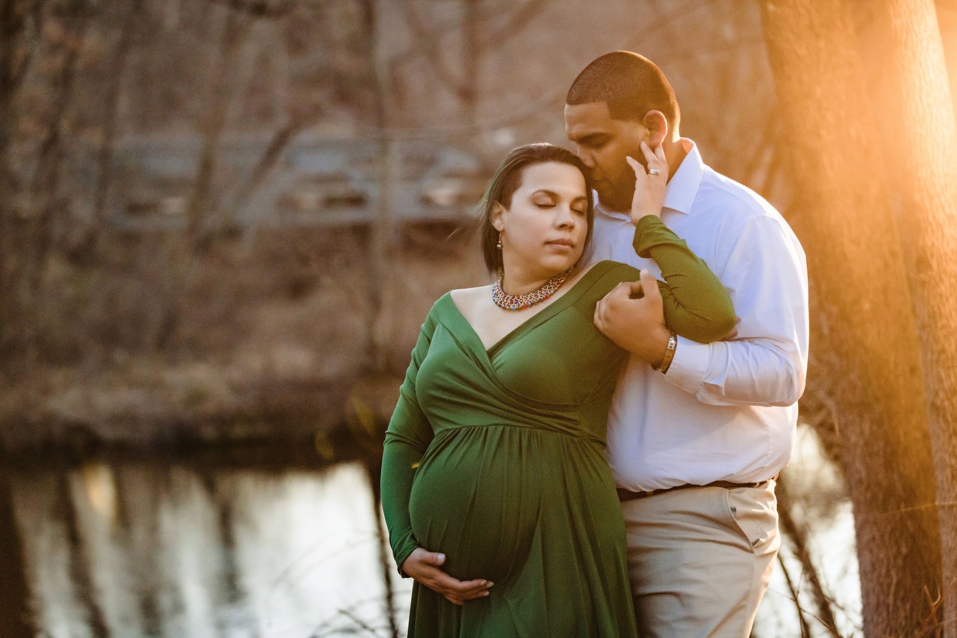 Couple by a lakeside at sunset, the woman in a green dress gently cradling her pregnant belly.