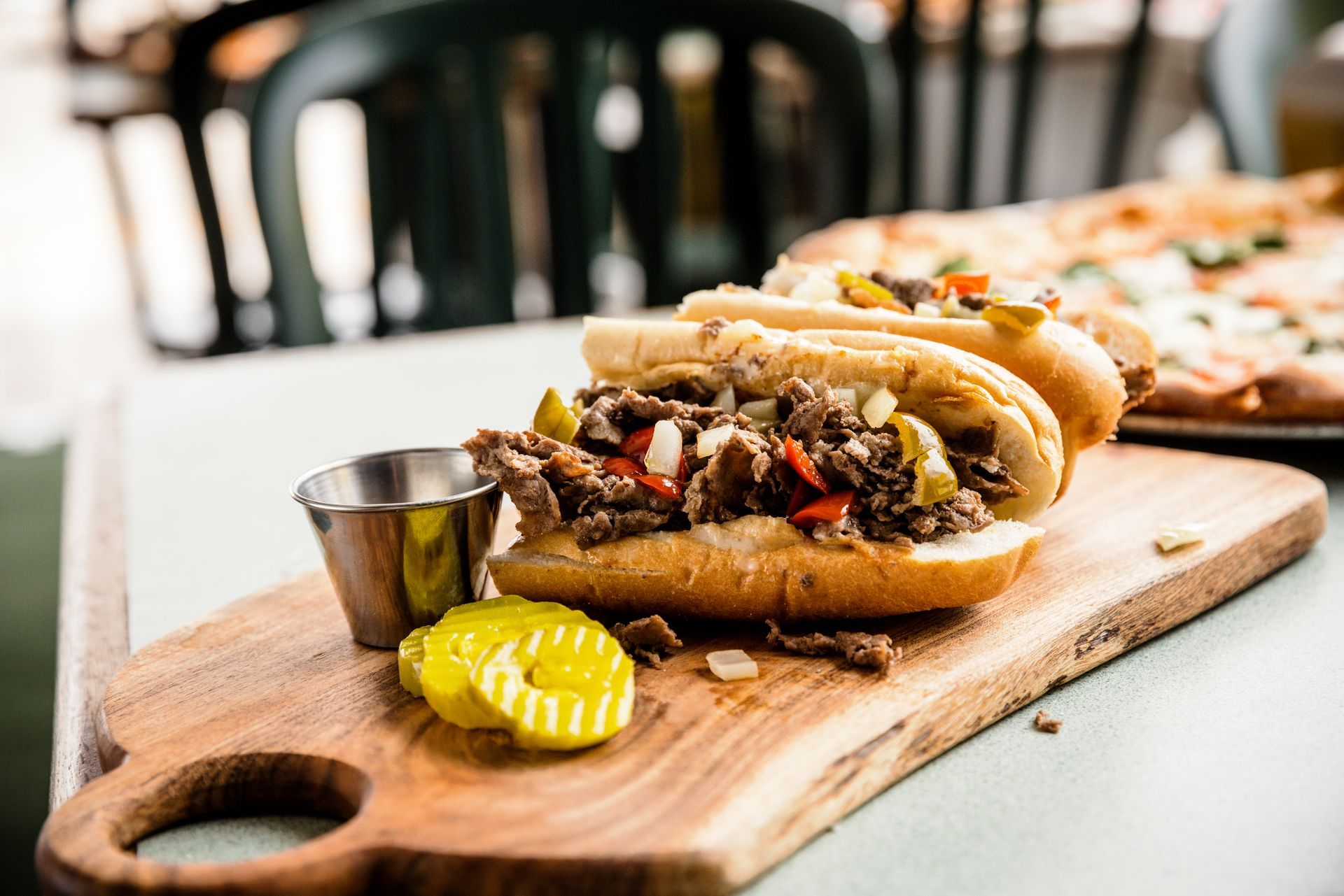 Hot dogs on a wooden board with pickles and a small metal sauce cup, served at a table.