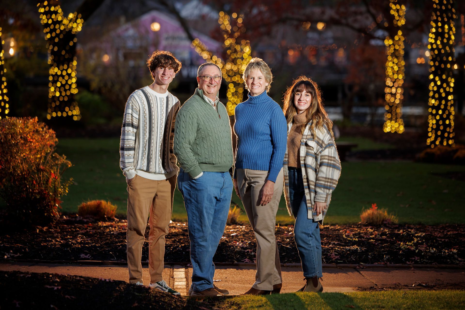 Four people posing outdoors at night near trees wrapped in lights, smiling in casual fall clothing.