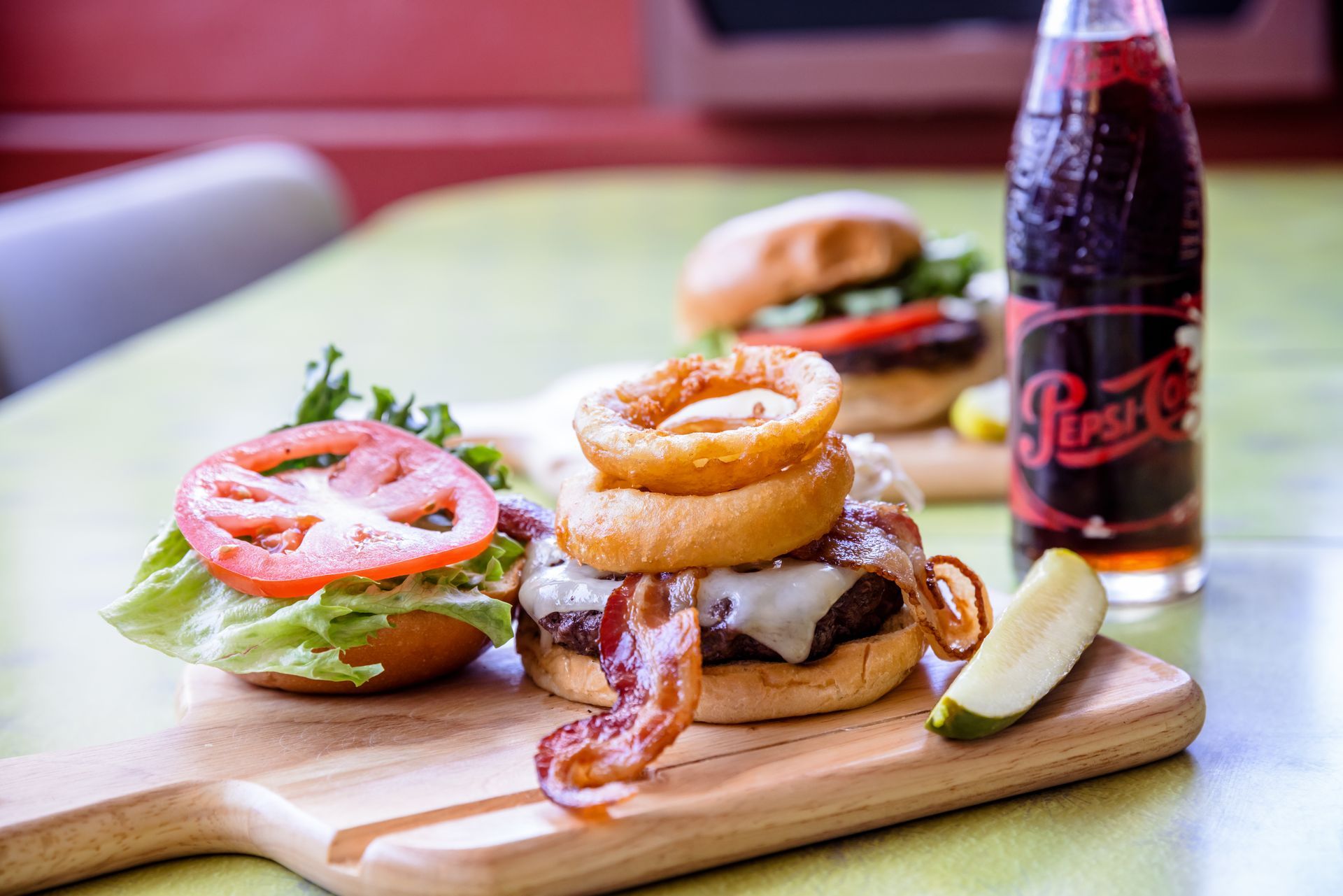 Burger with onion rings, tomato, lettuce, fries, pickle, and a soda bottle on a wooden board