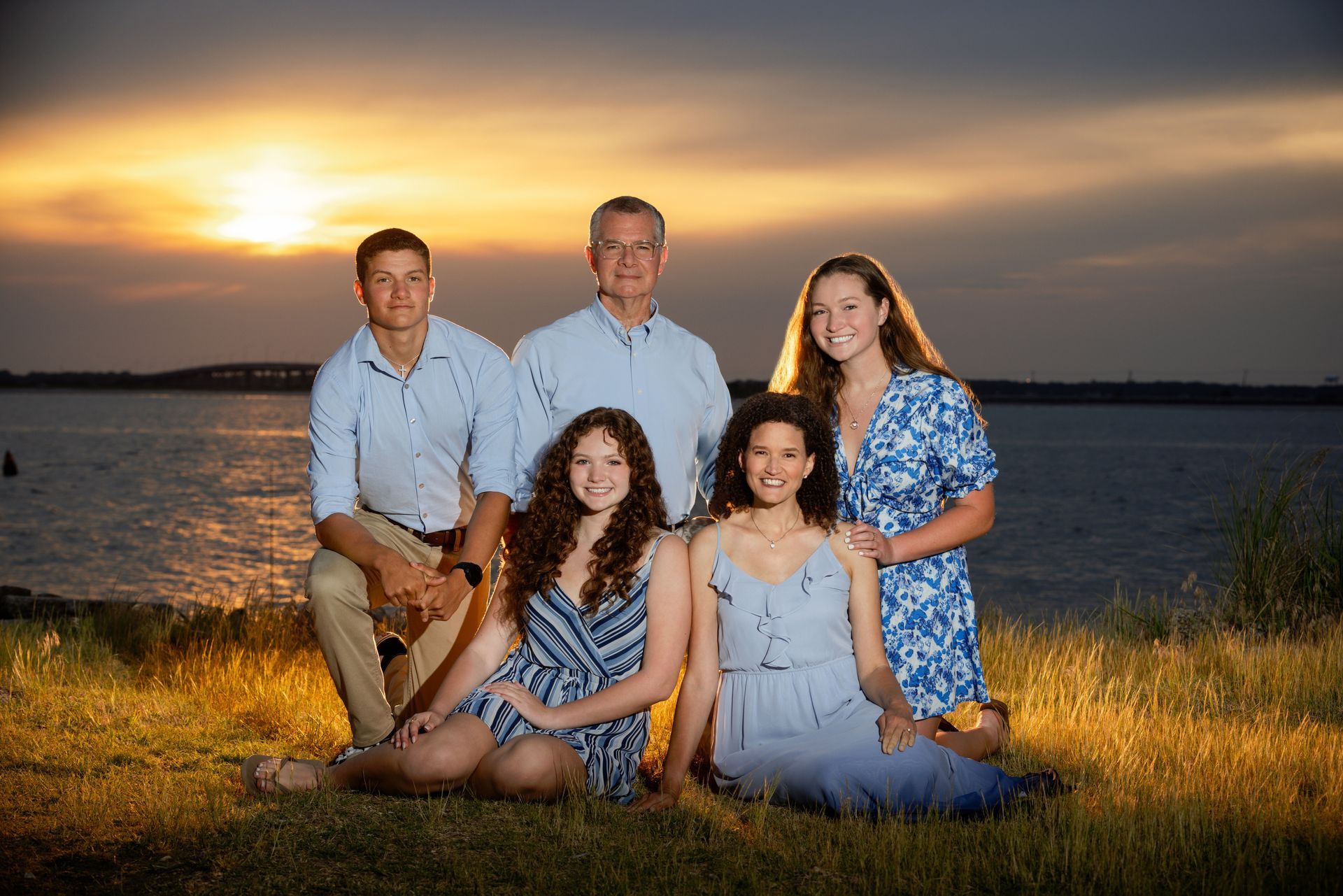 Five people posing outdoors by a lake at sunset, seated and standing in the grass.