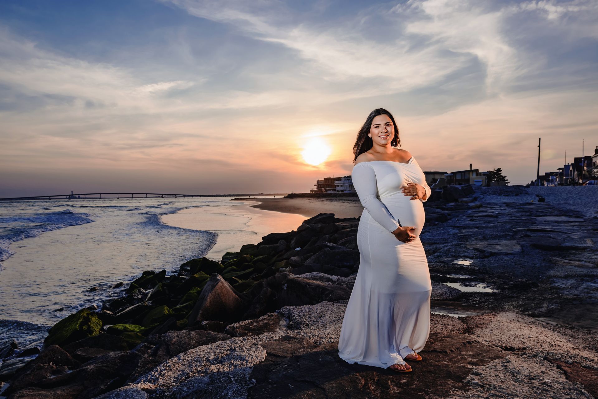 Pregnant woman in a white dress standing on rocks by the ocean at sunset