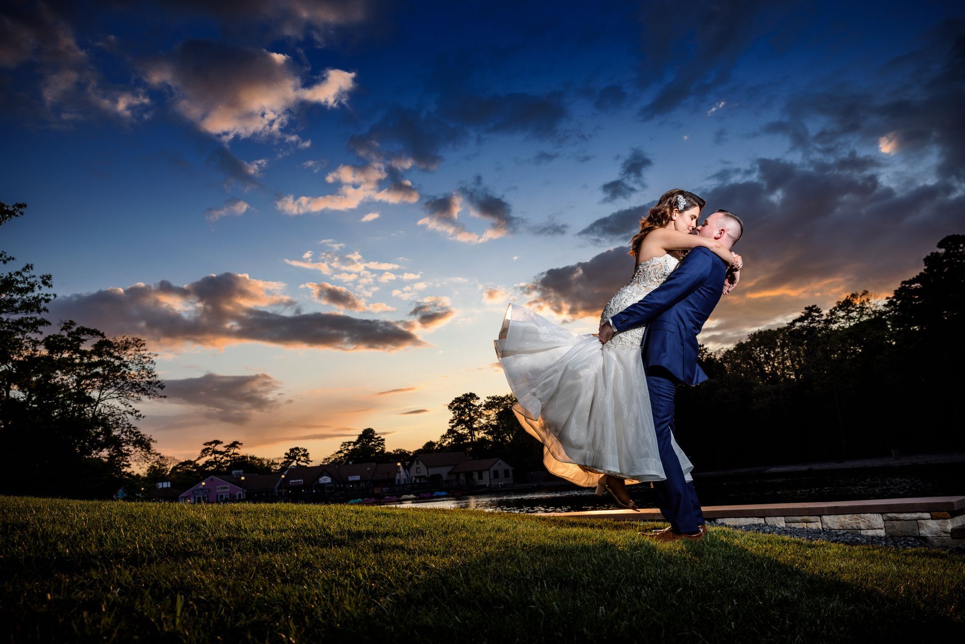 Couple embracing in a field at sunset, with a dramatic cloudy sky and trees silhouetted behind them.